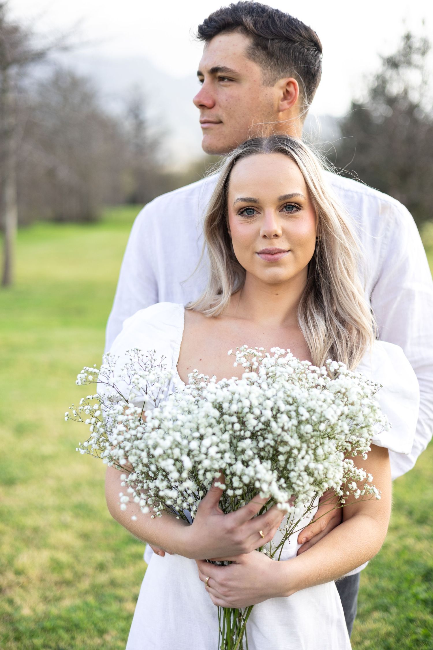 Couple in white wedding attire poses with delicate baby's breath bouquet in outdoor spring setting.