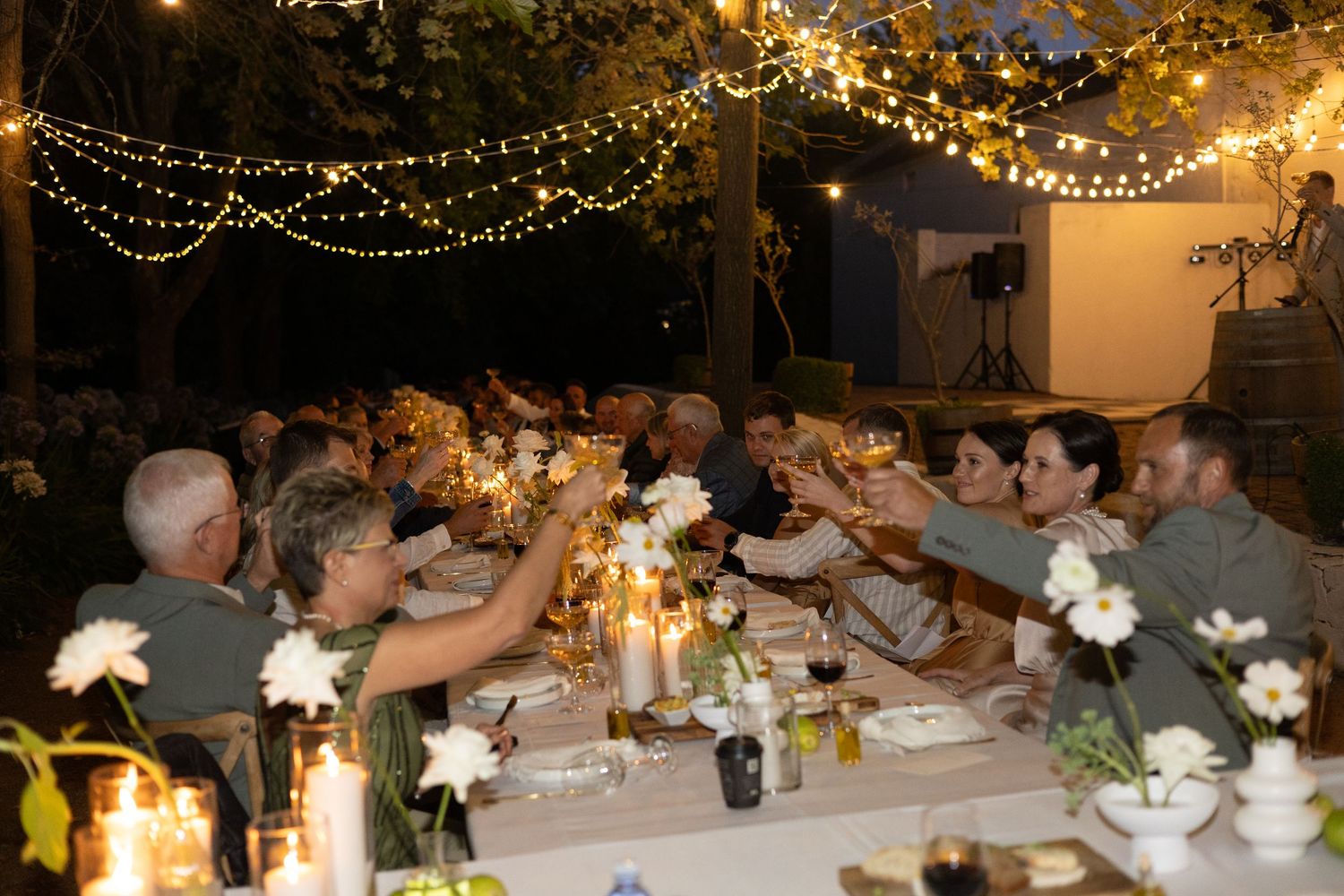 People toast and celebrate at an outdoor dinner party under twinkling string lights and candles at dusk.