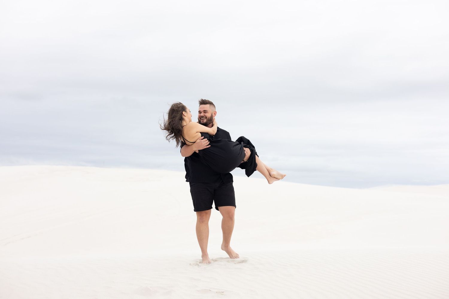 Person carrying partner while standing barefoot on white sand beach.