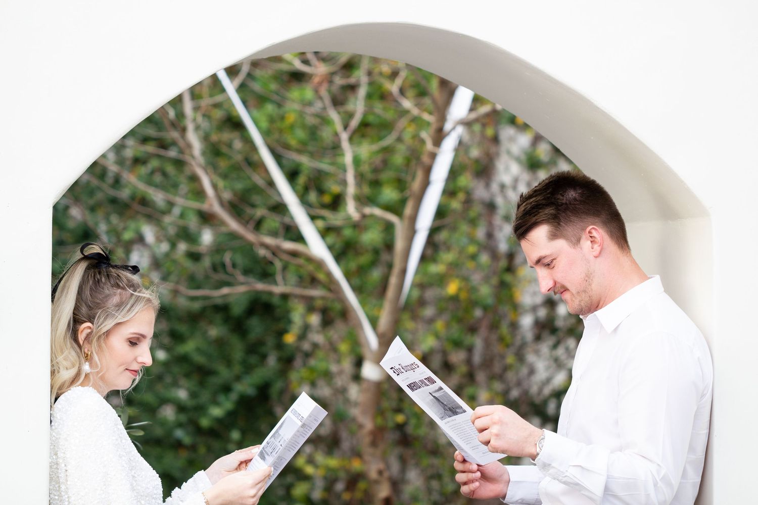 Two people reading documents while standing in an outdoor archway surrounded by greenery.