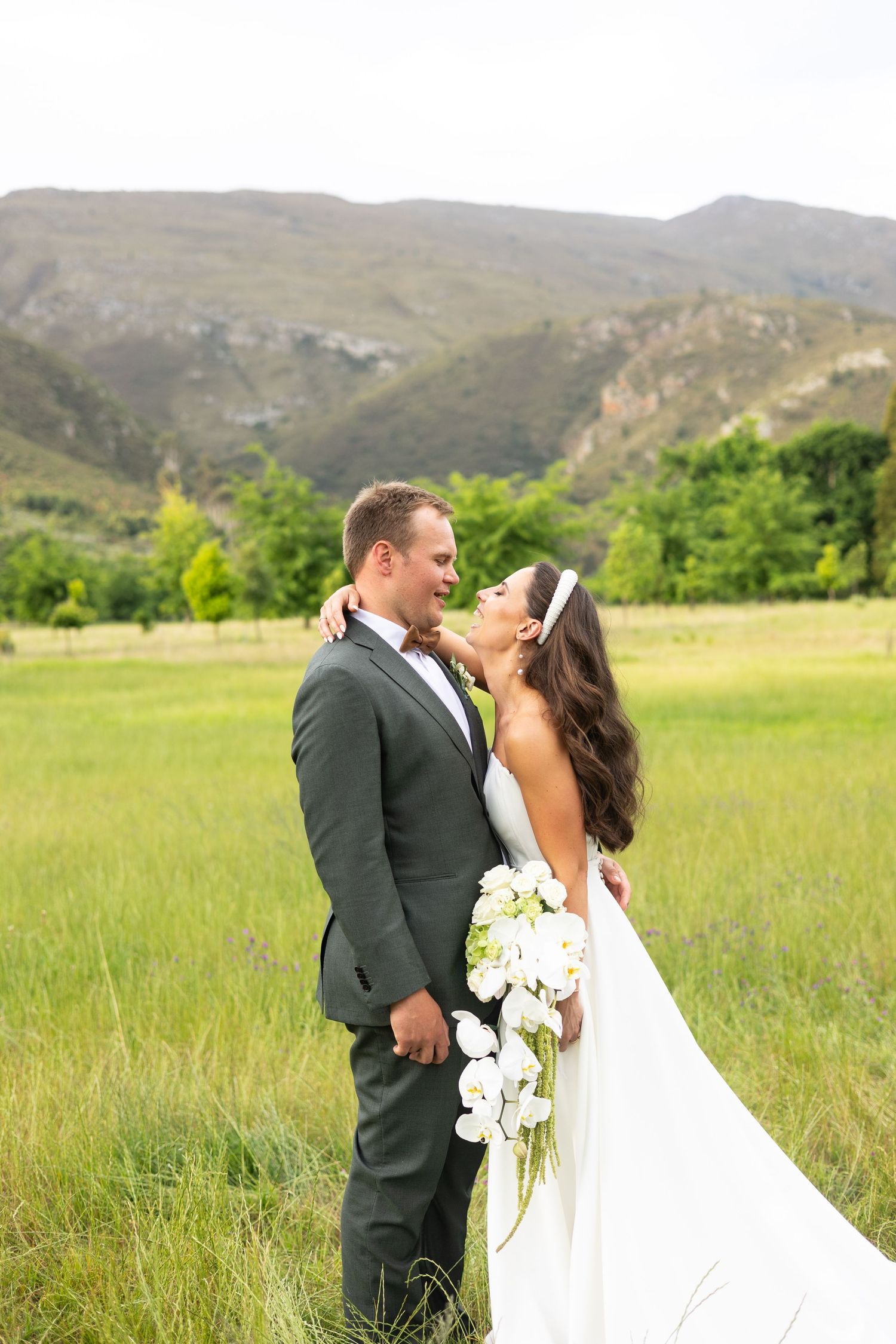 Couple in wedding attire embrace in a scenic mountain meadow with flowing white gown and grey suit.