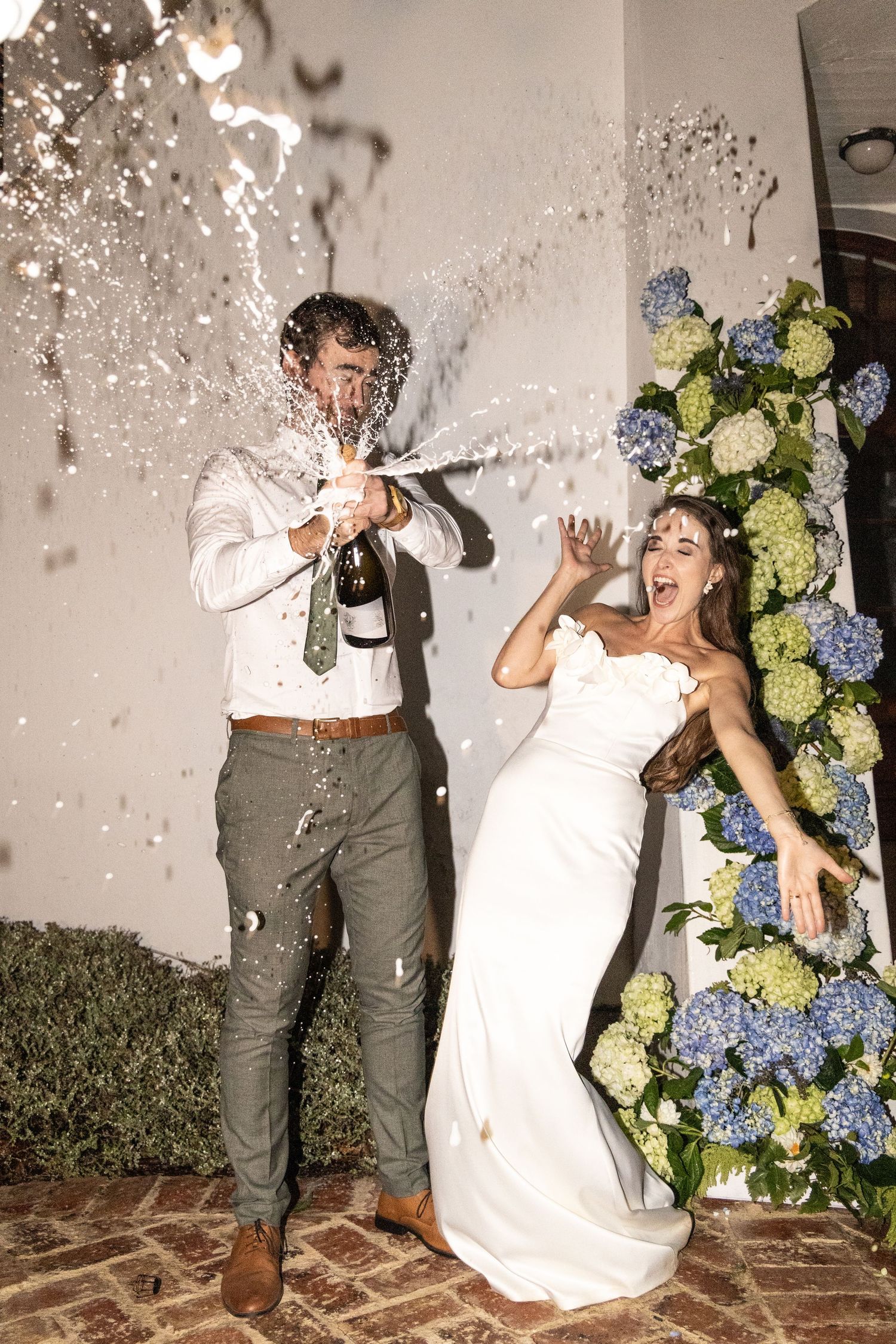 A sequence of playful wedding photos shows a couple popping and spraying champagne against a white wall with blue hydrangeas.