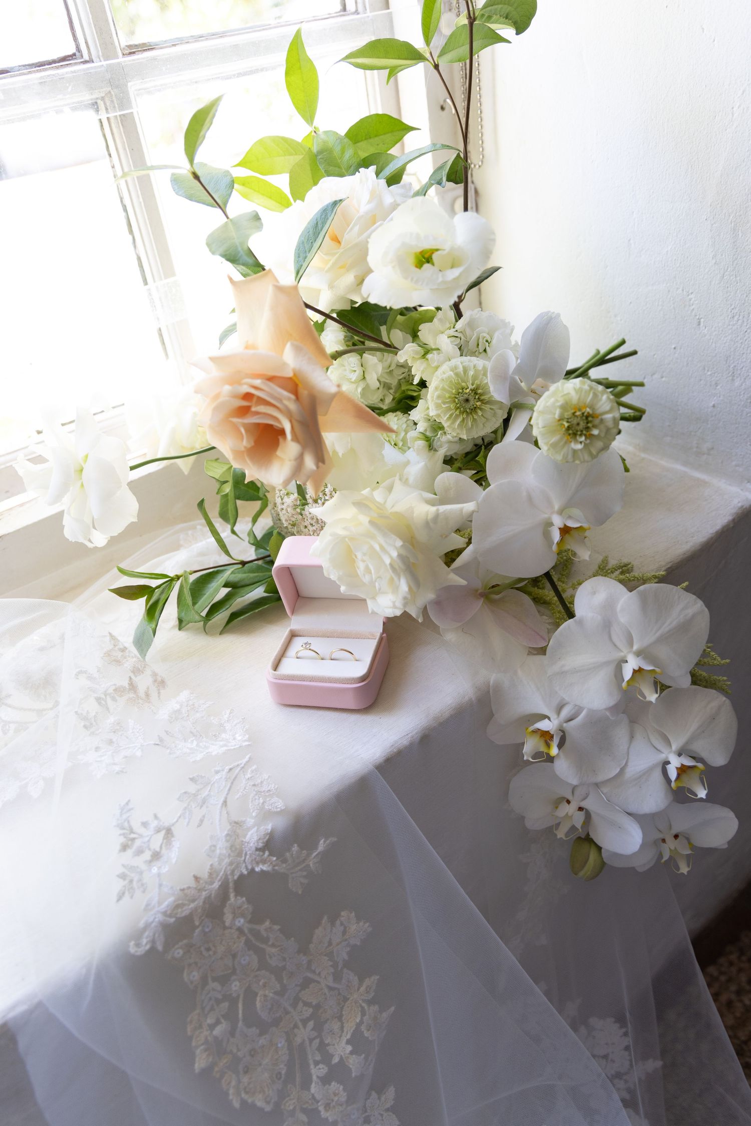 Elegant bridal bouquet of white orchids and roses with ring box on lace fabric.