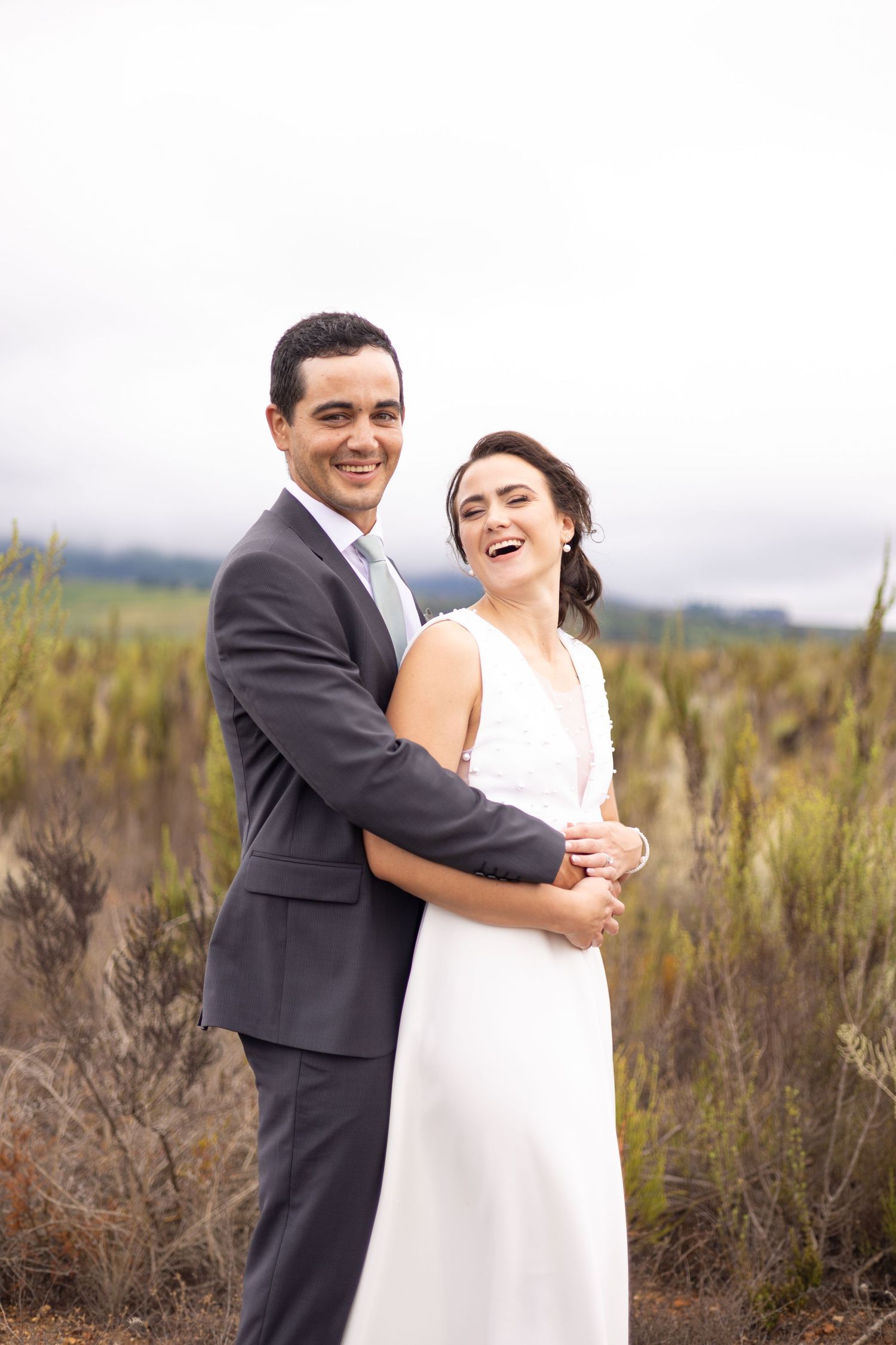 A couple in wedding attire embrace while laughing together in a natural outdoor setting with tall grass and overcast skies.