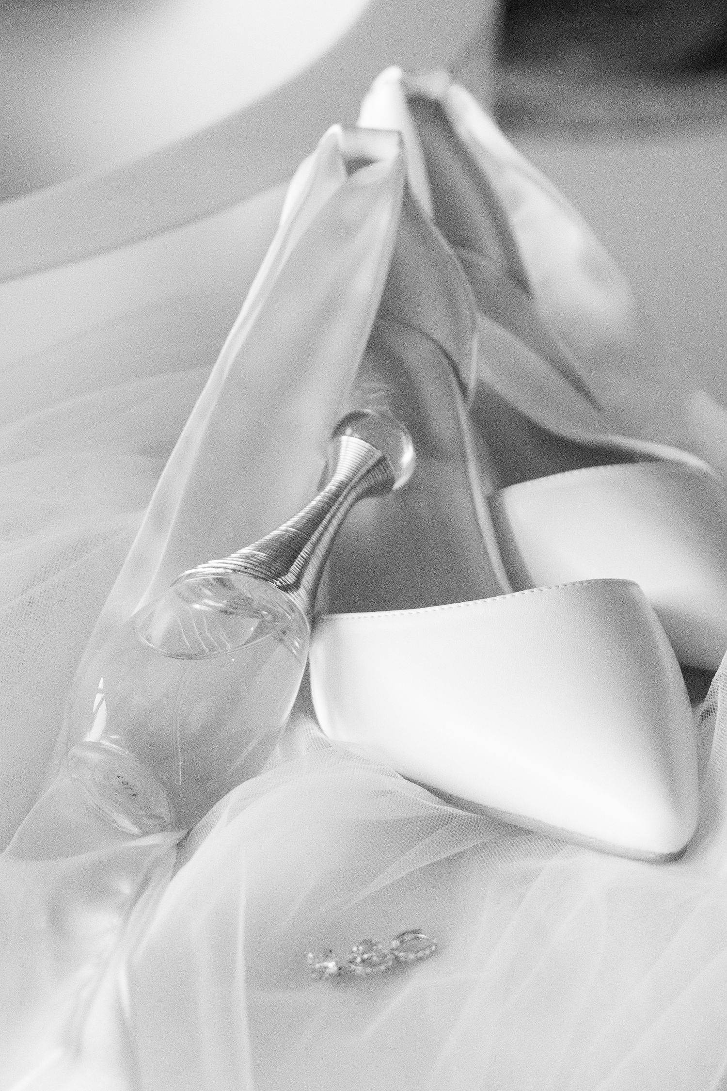 Wedding accessories including white bridal shoe, veil, and engagement ring captured in elegant black and white photography.