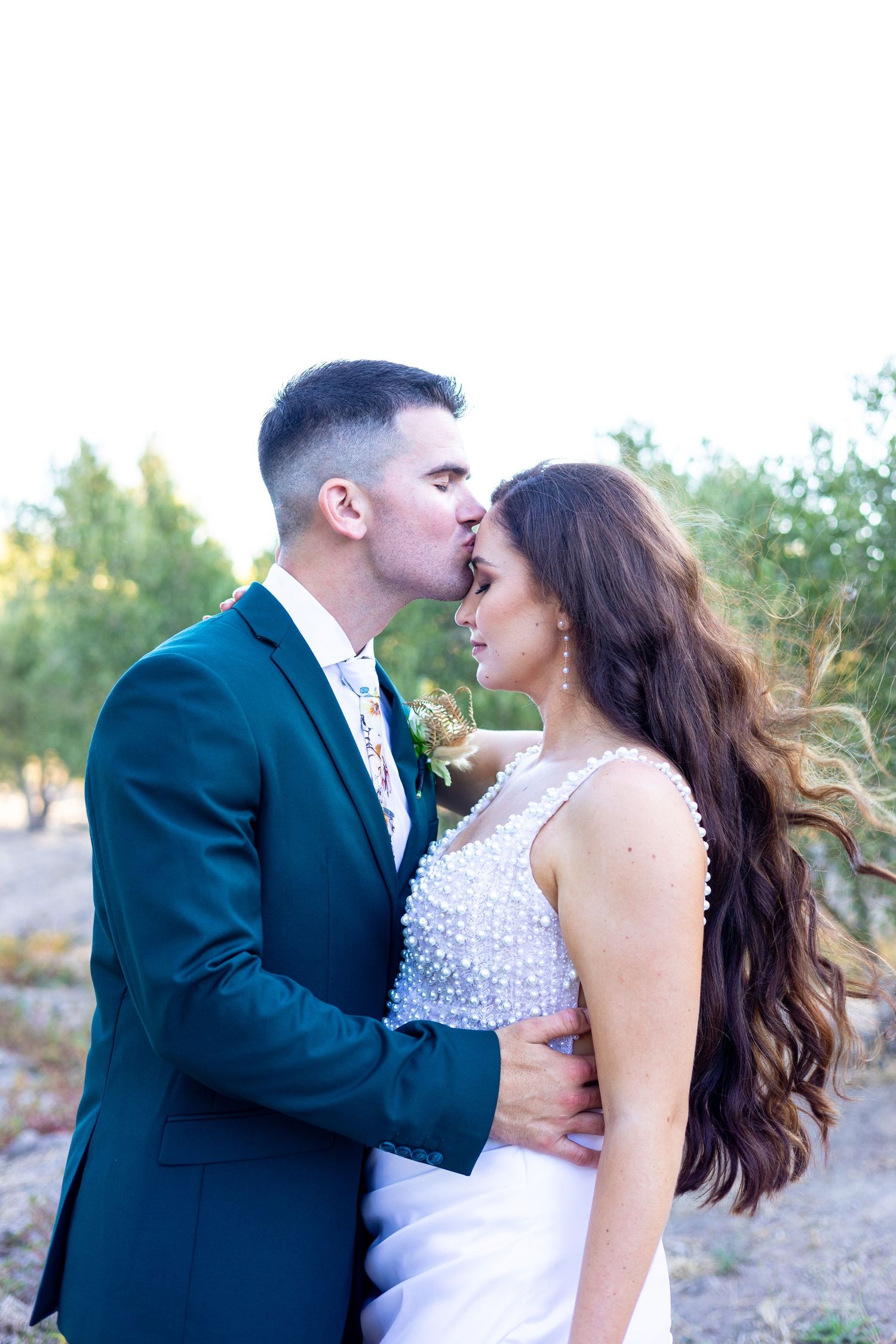 A romantic wedding photo featuring a tender moment between bride and groom outdoors at sunset with greenery in background.