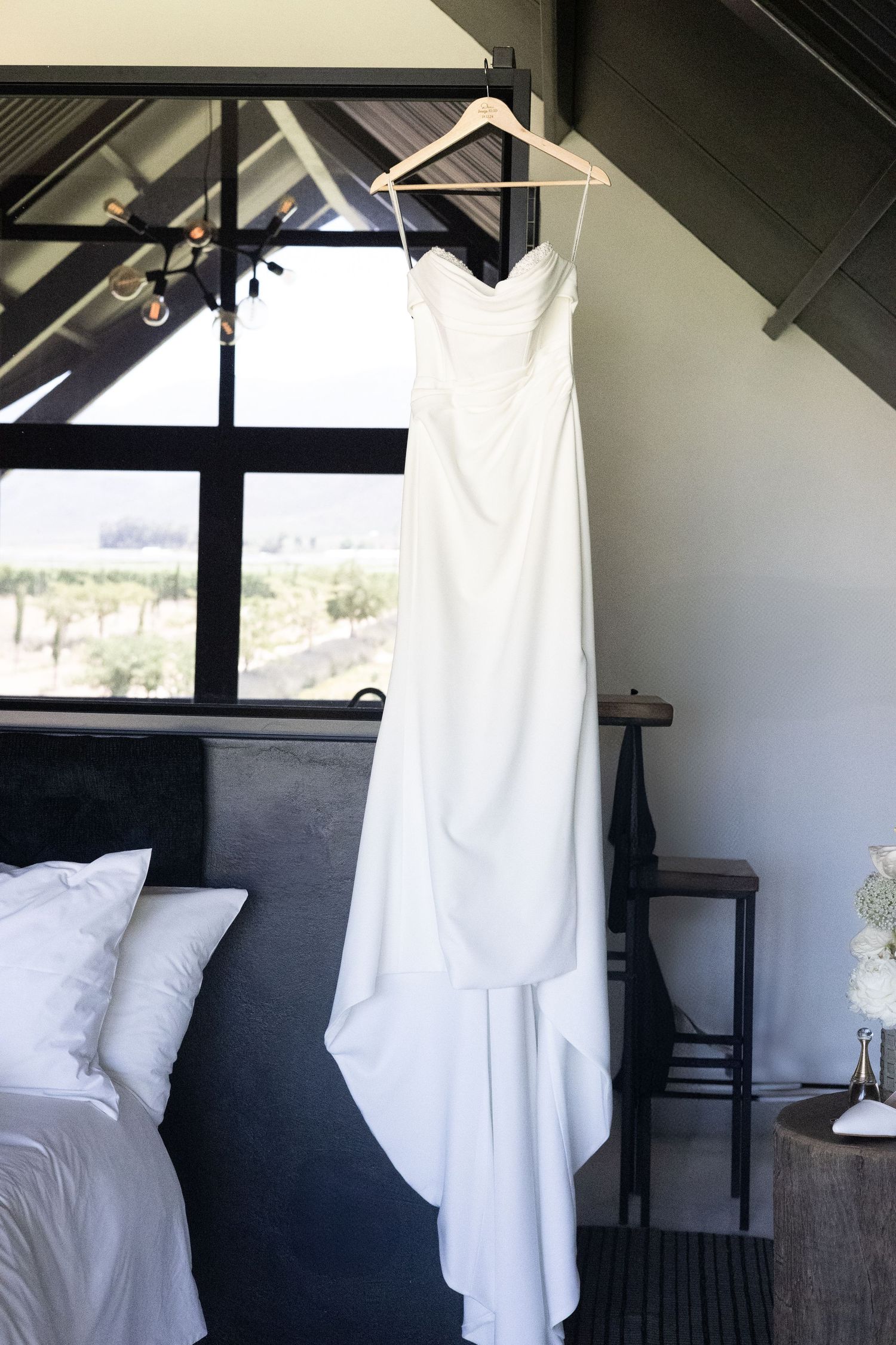 A white wedding dress hangs from a wooden hanger against a dark window frame in a bedroom with white bedding.