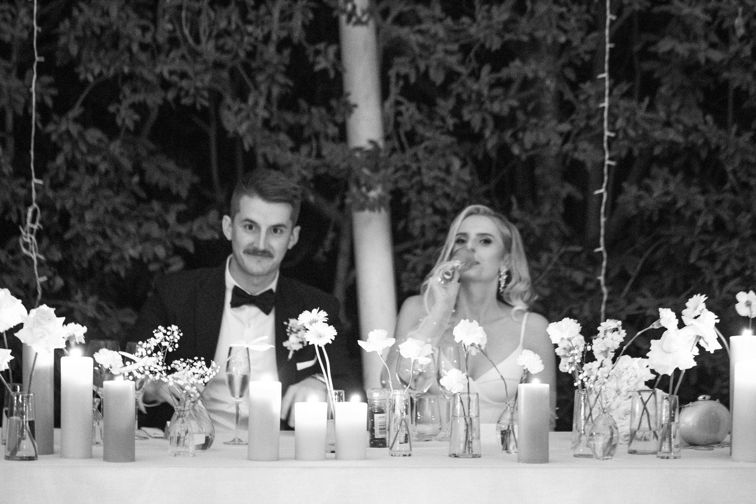 Wedding couple seated at elegant reception table with candles and floral centerpieces.