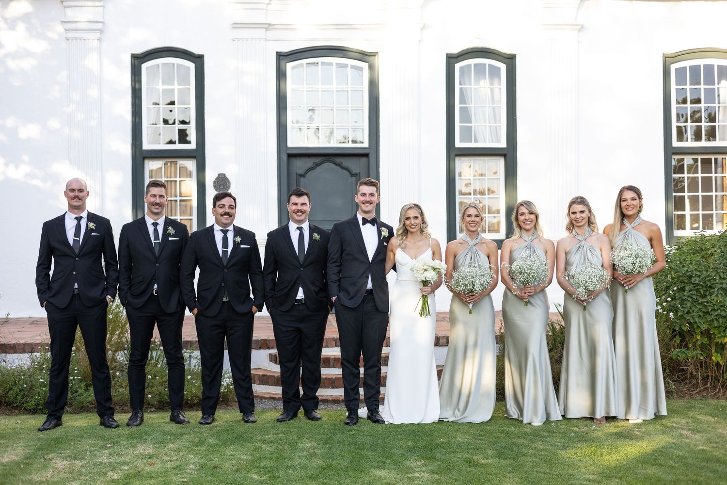 Wedding party in formal attire lined up against white colonial building with black windows.