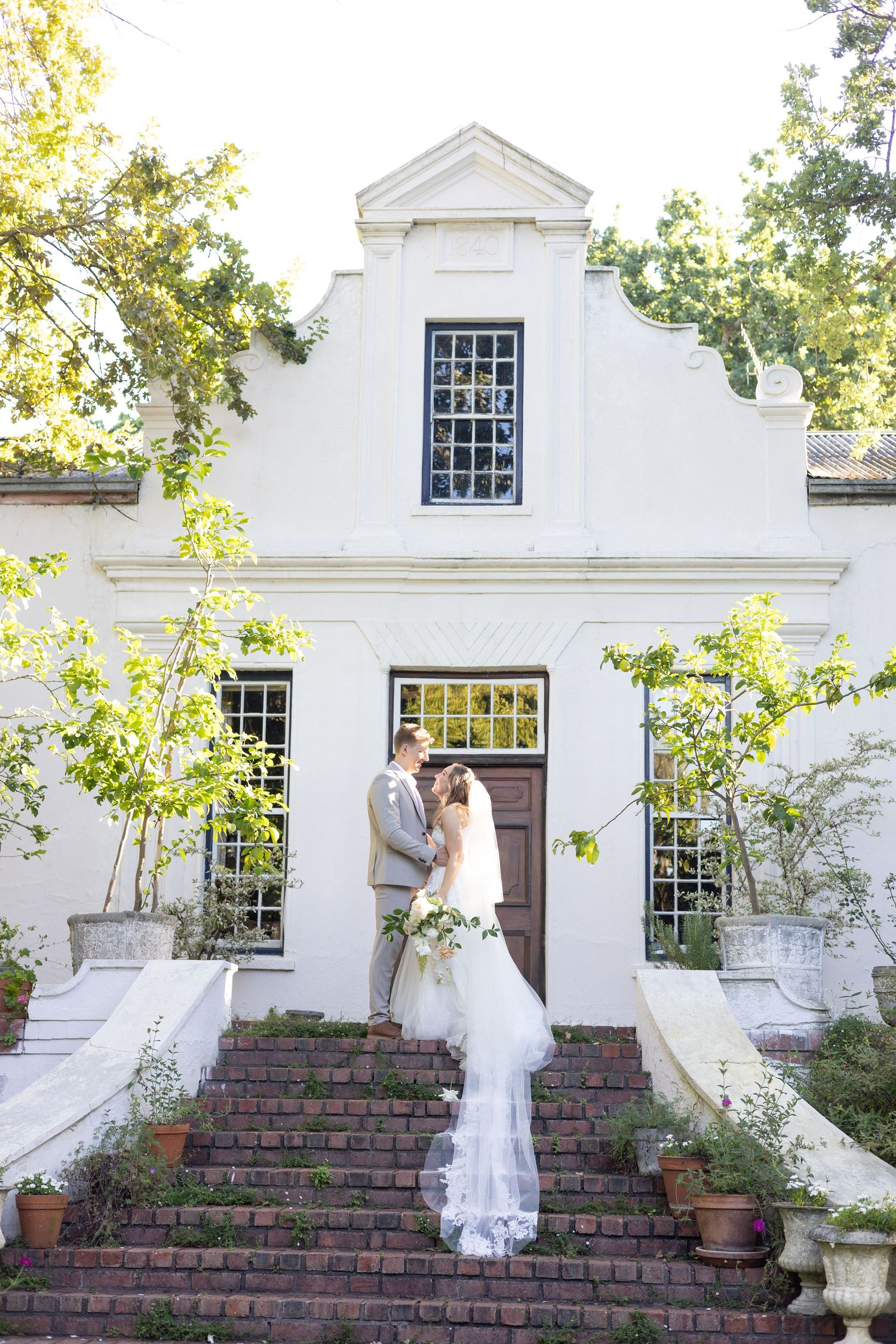 Wedding couple standing on brick steps of a white colonial-style building with ornate windows.