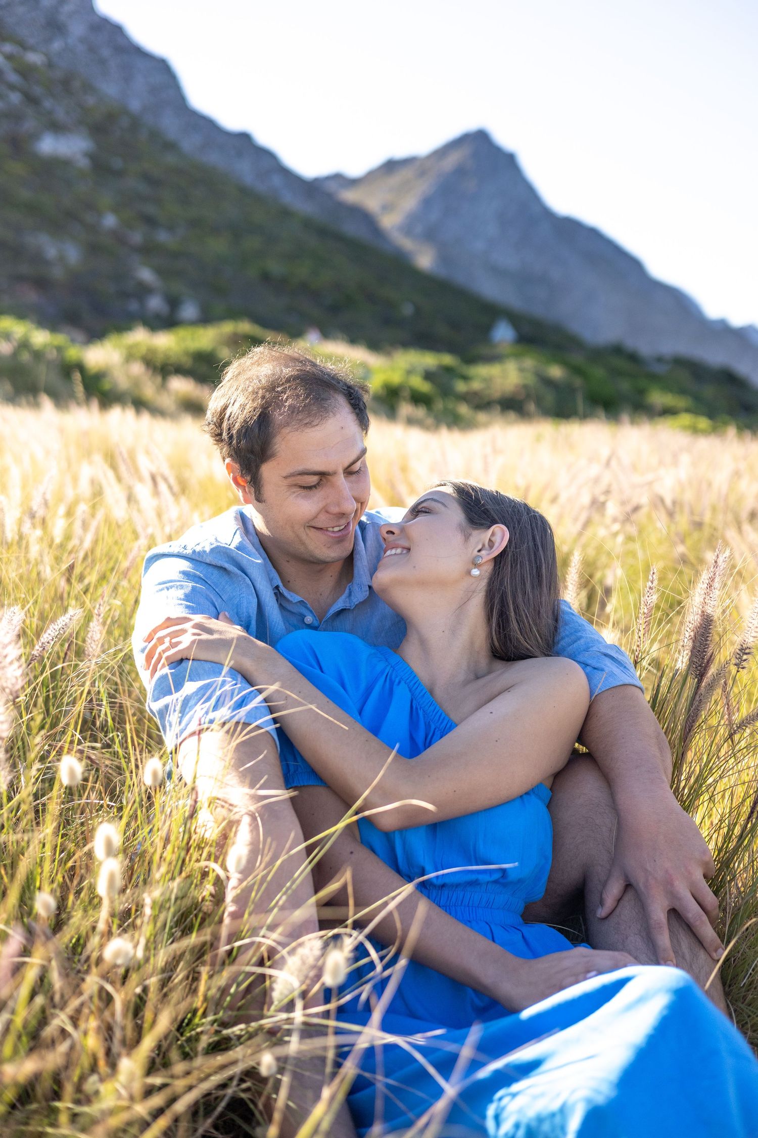 A couple embracing and laughing while sitting in a golden field with mountains in the background.