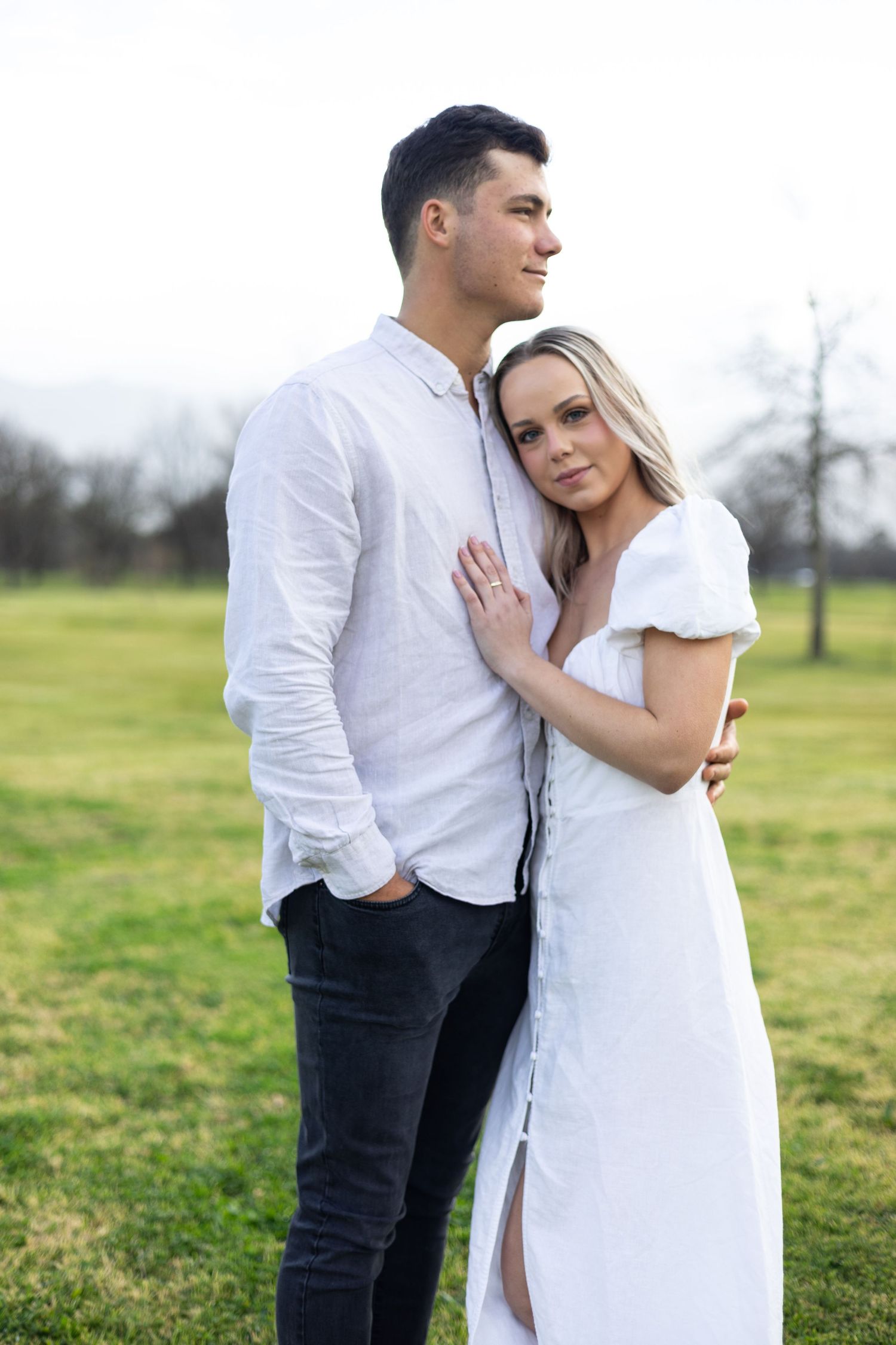 Couple in white clothing embracing in meadow on cloudy day.