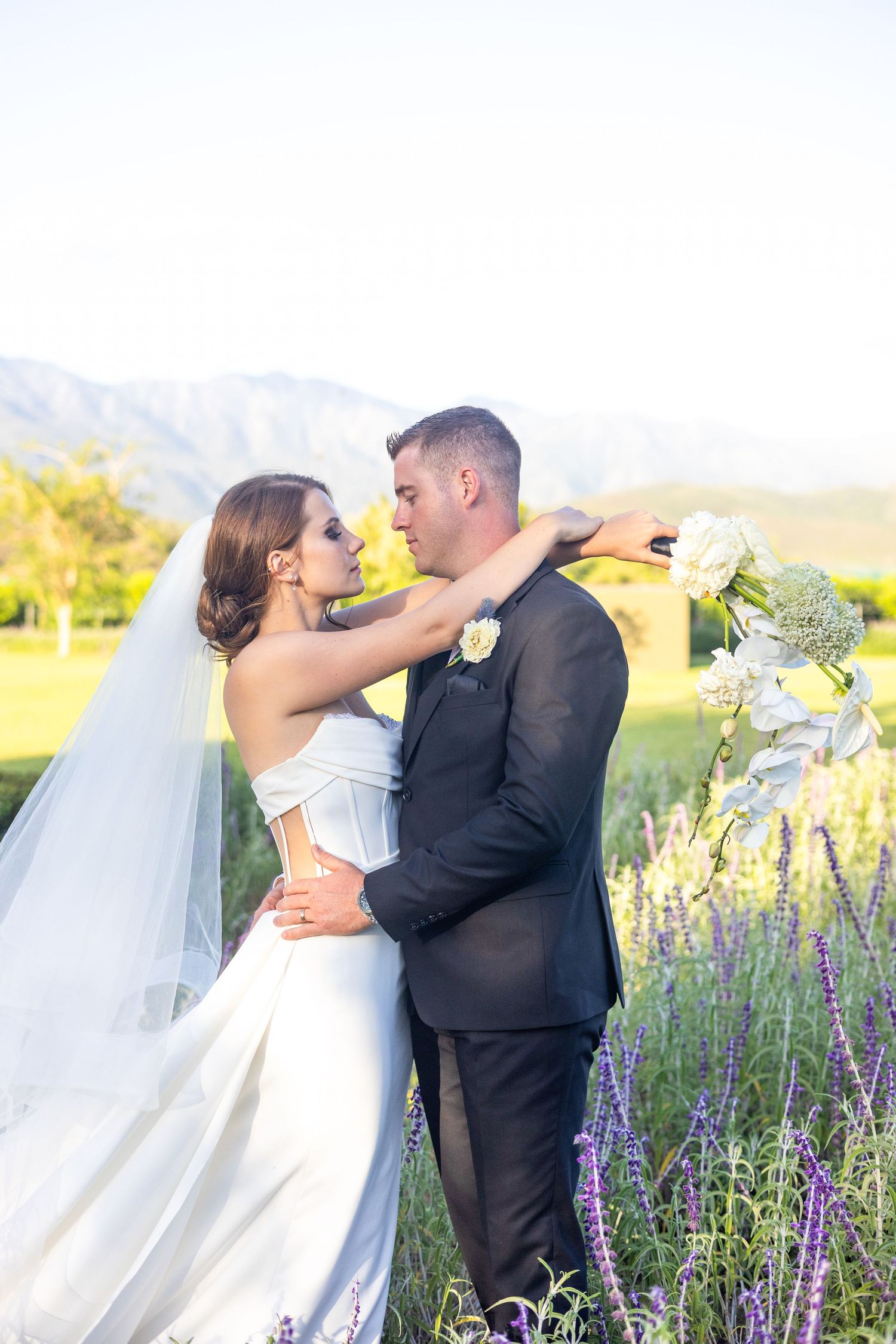 Newlywed couple embracing in field of lavender with mountains in background during their outdoor wedding.