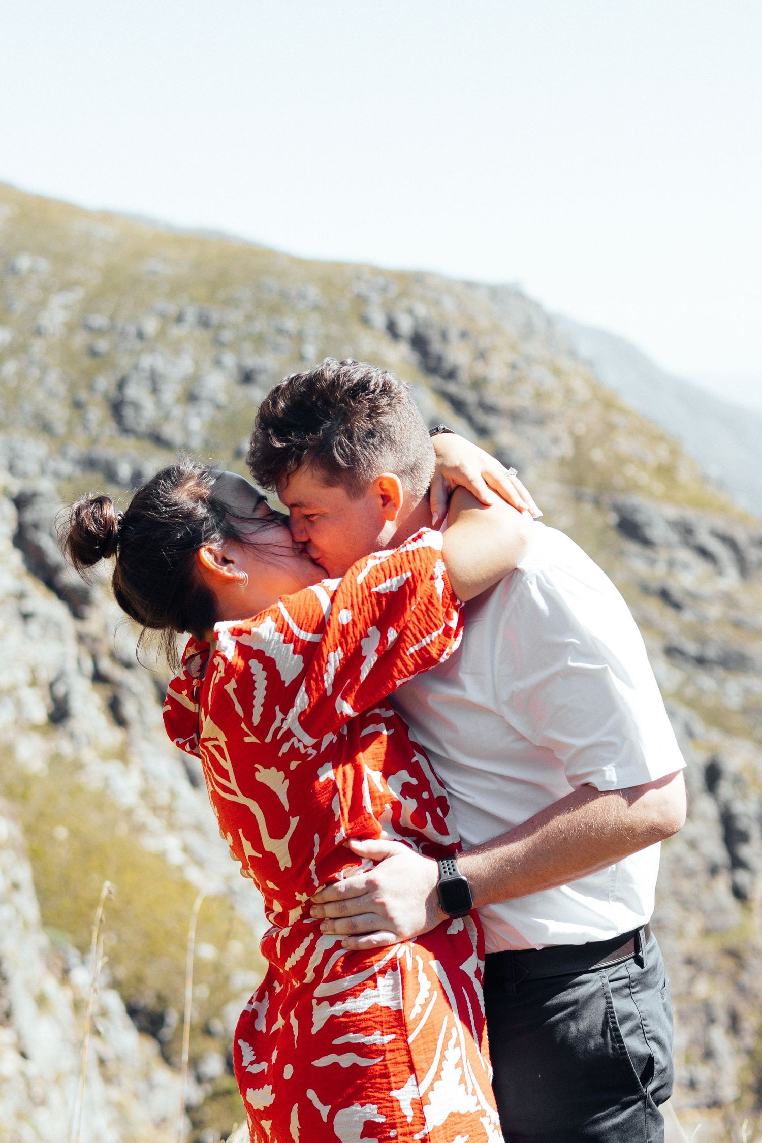 A couple embraces against a mountain backdrop, with one wearing a vibrant red patterned dress.