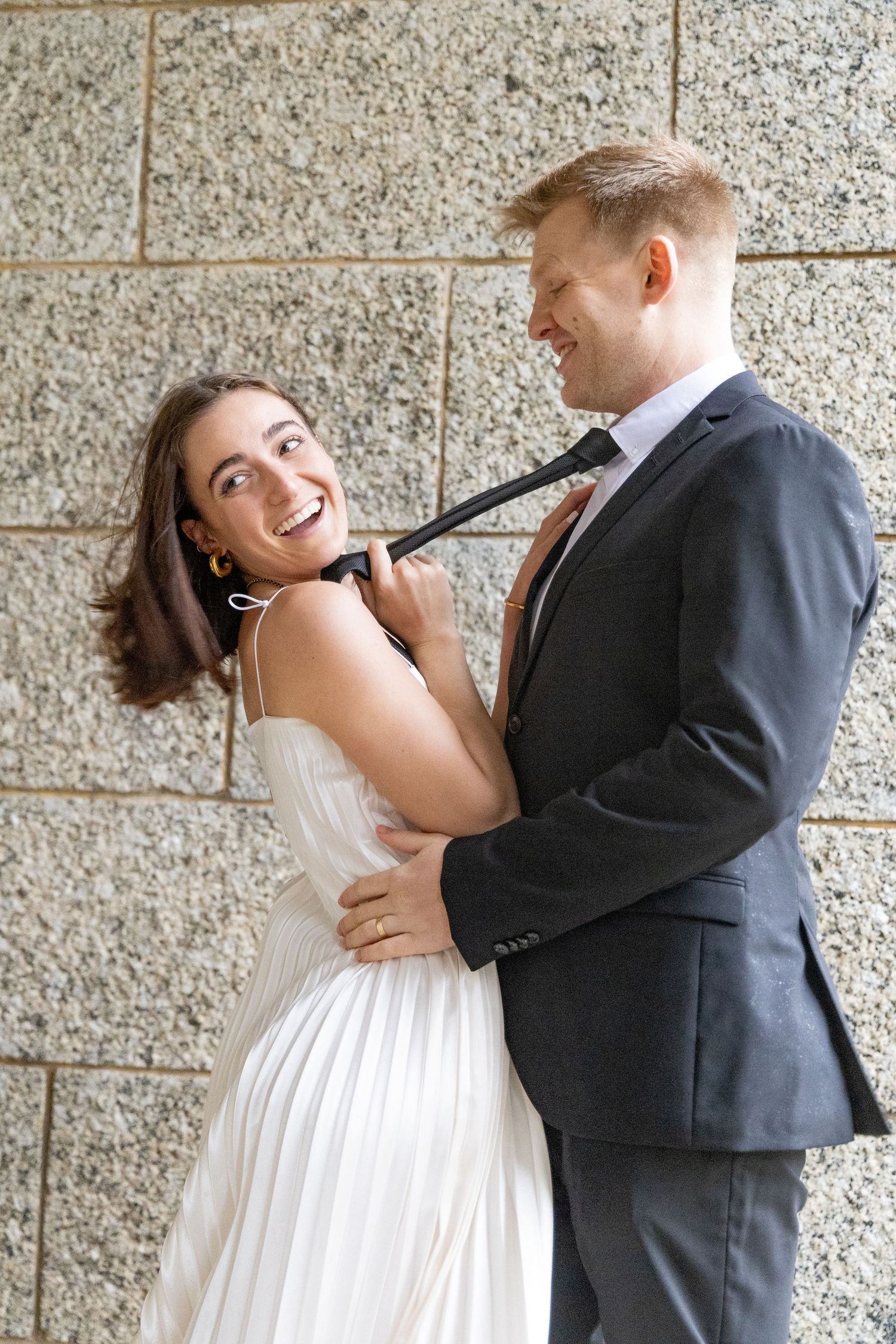Playful couple in formal attire laughing against stone wall.