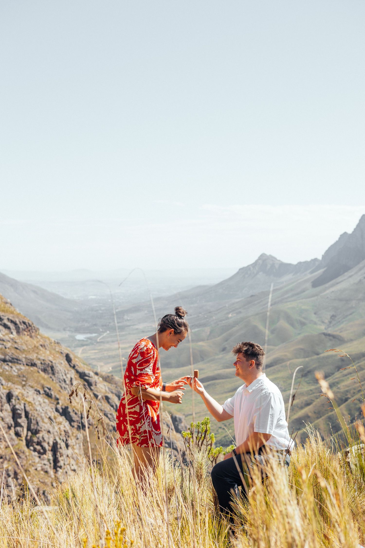 A scenic mountain proposal moment with a couple standing among tall grass on a hillside.
