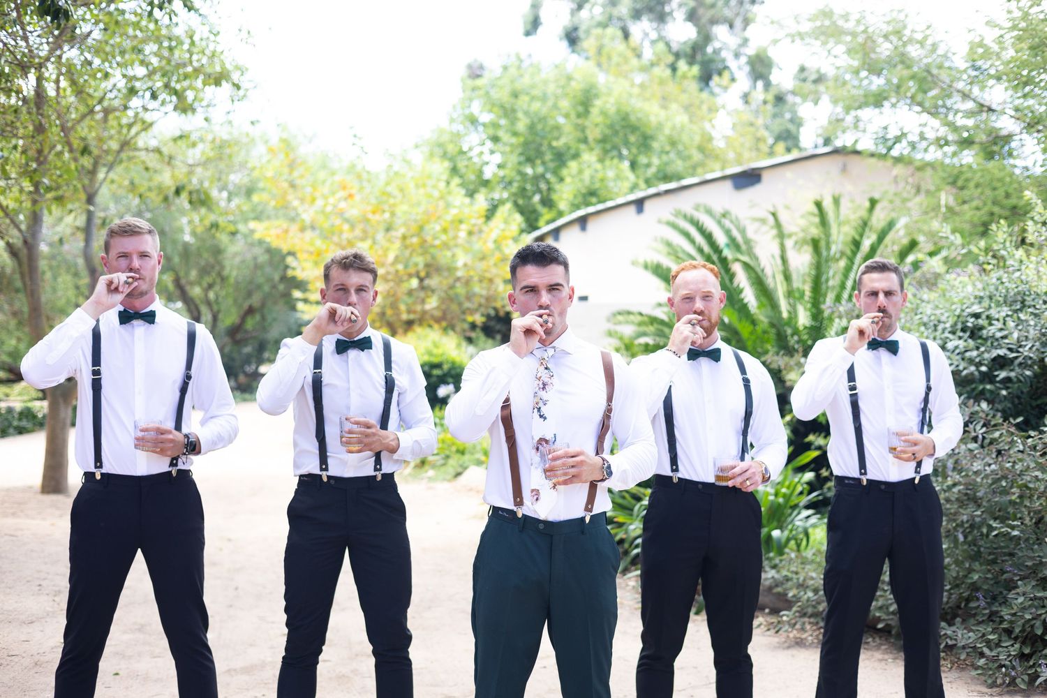 Groomsmen in black pants, white shirts and suspenders pose outdoors in a garden setting.