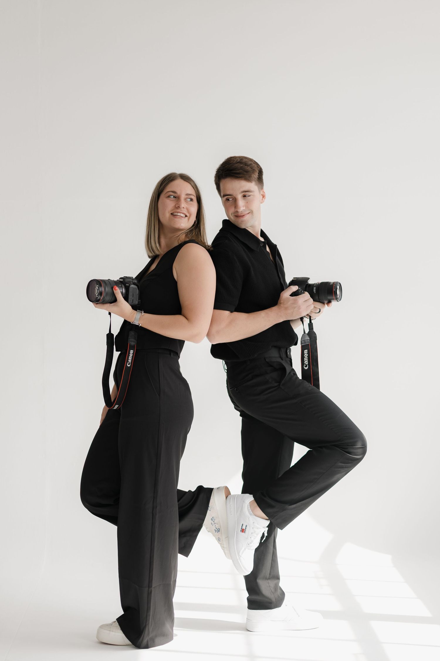 Two photographers in matching black outfits pose back-to-back with cameras against a white studio background.