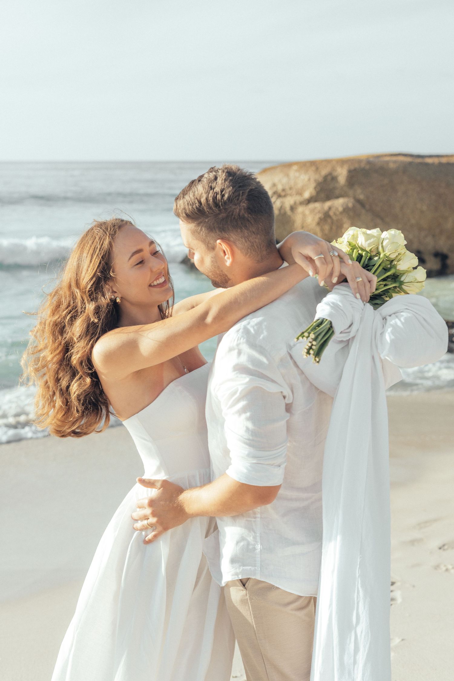 A romantic beachside moment with a couple in white attire embracing and holding a bouquet against a coastal backdrop.