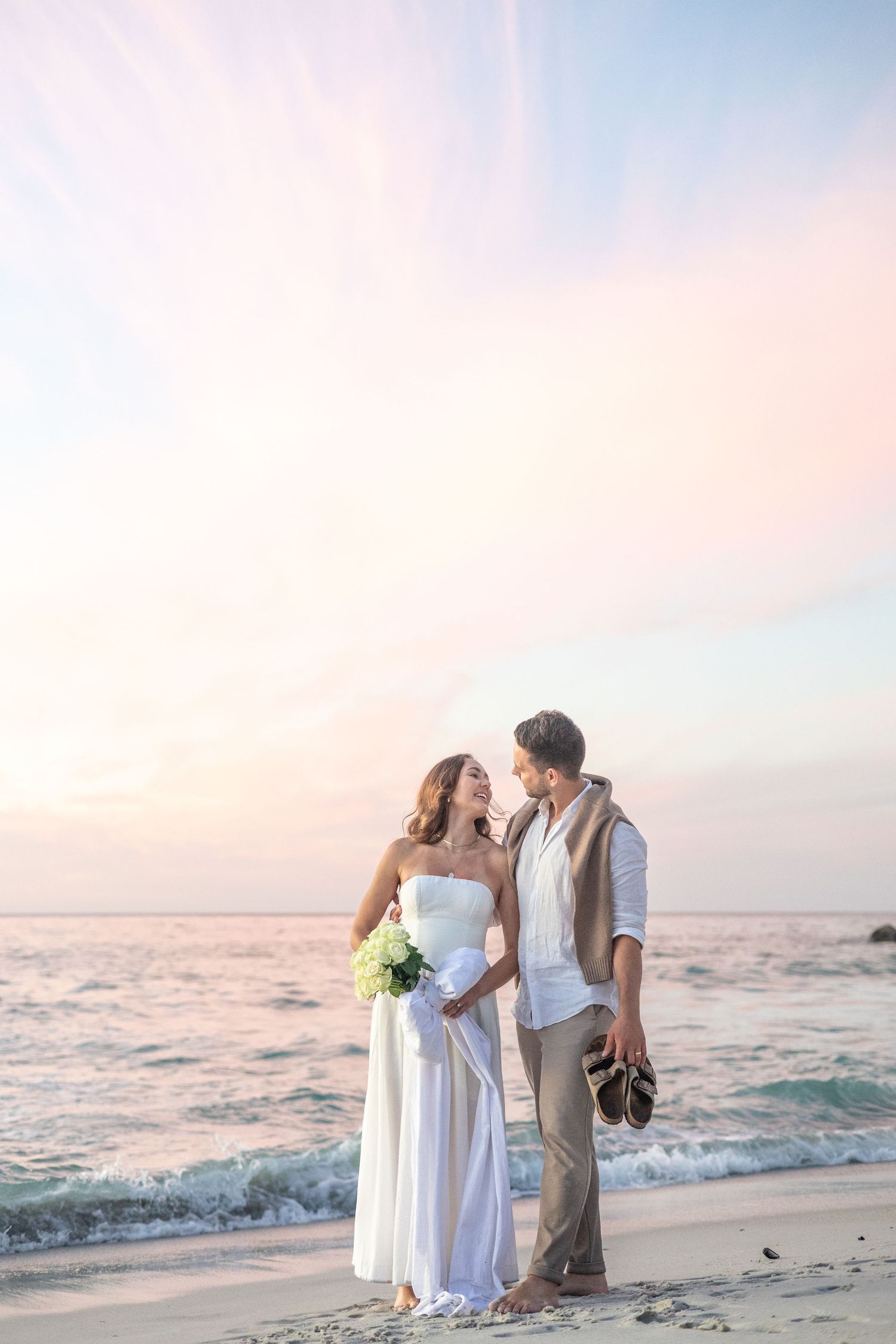 Couple stands together on beach at sunset holding bouquet with ocean waves behind them.