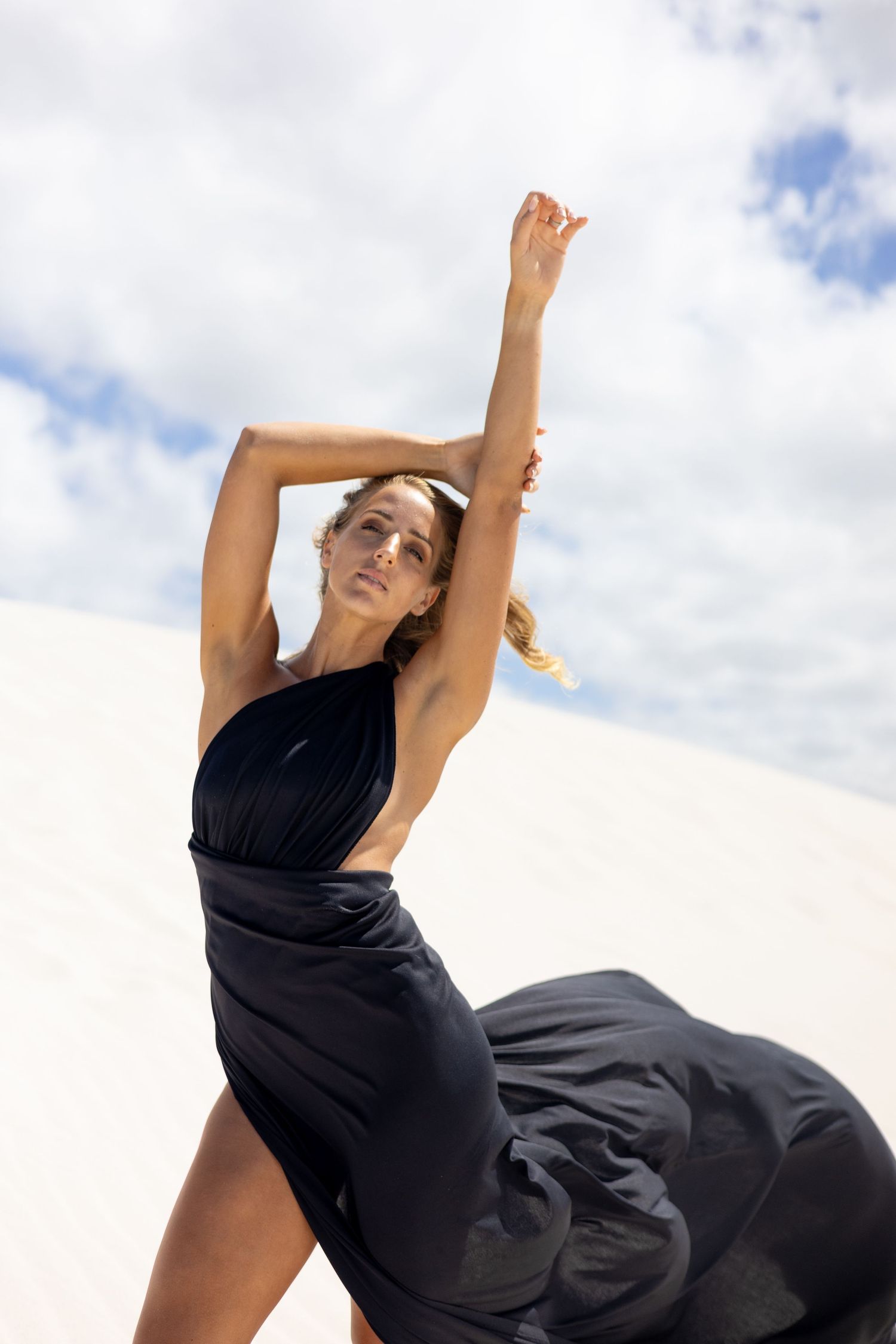 Woman in flowing black dress poses gracefully against cloudy sky in outdoor photoshoot.