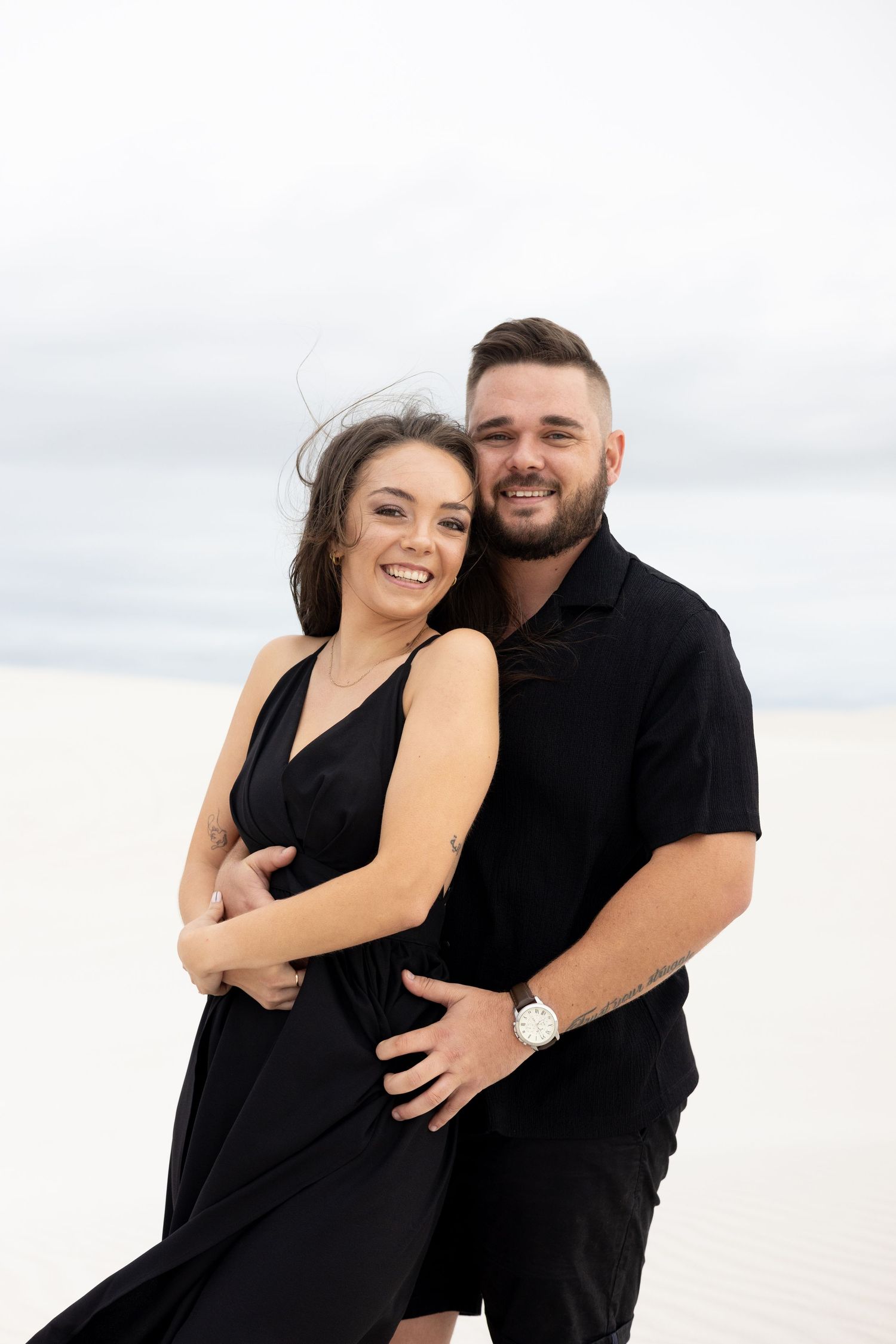 A couple in matching black outfits poses together on a bright white sandy beach.