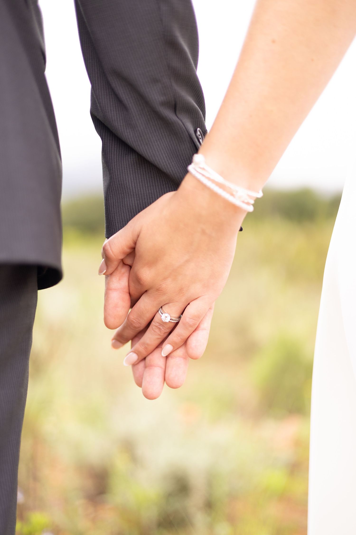 Close-up of newlyweds holding hands showing wedding rings and bracelets against blurred outdoor background.