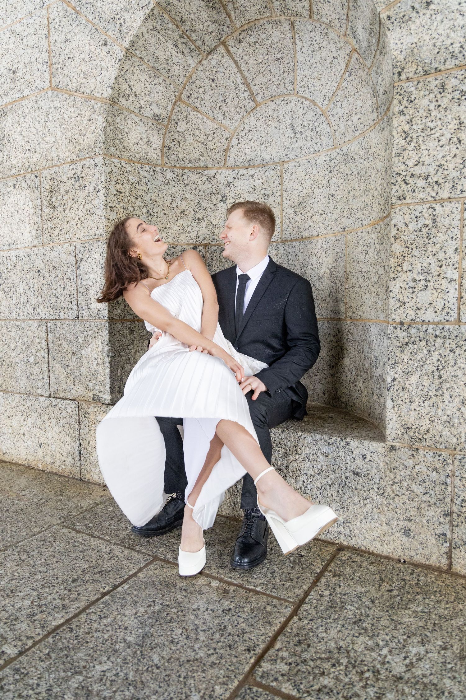 Playful wedding couple poses against stone archway in romantic moment.