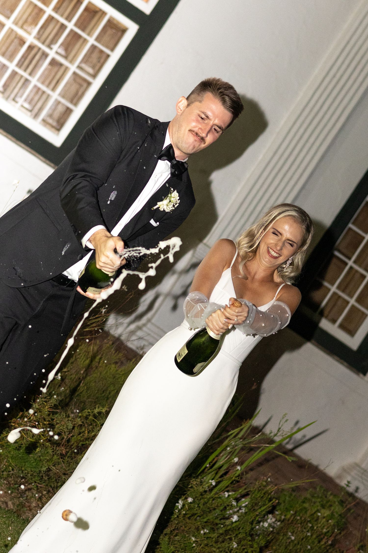 Joyful couple celebrates with champagne spray outside during wedding reception.