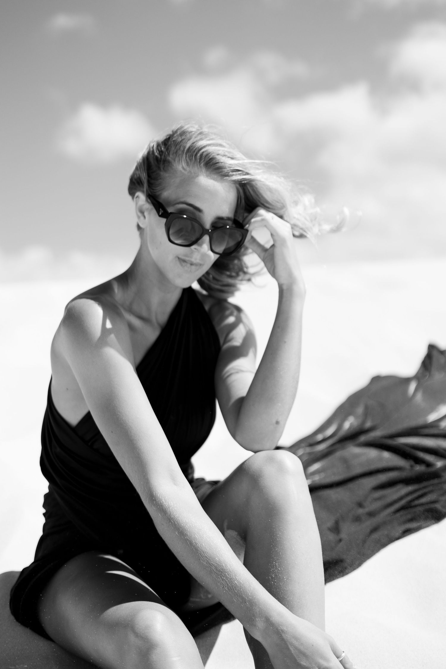 Woman in black swimsuit and sunglasses poses against bright sky in artistic black and white photo.