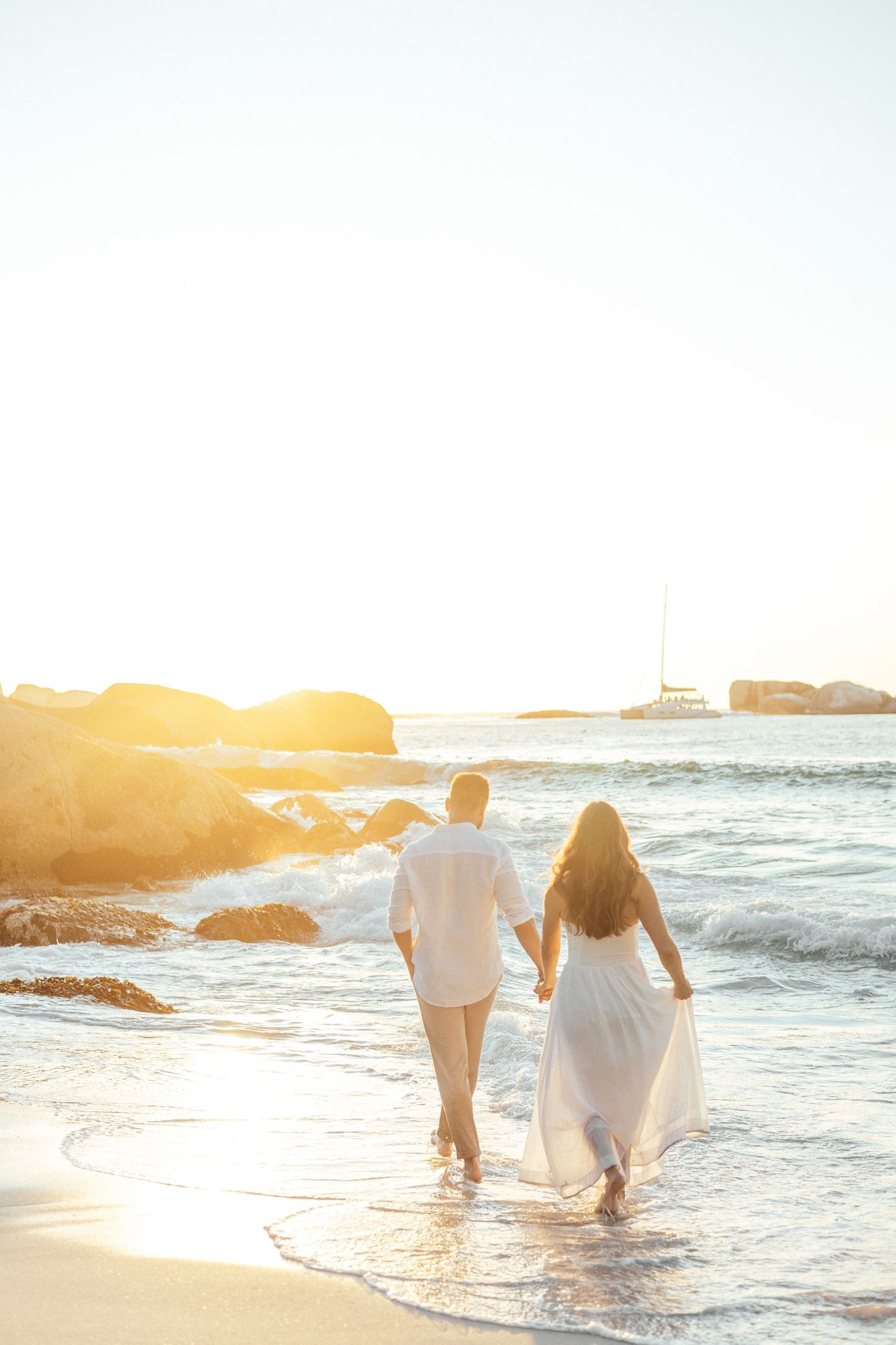 Couple walks hand in hand on beach at sunset with boat visible in distance.