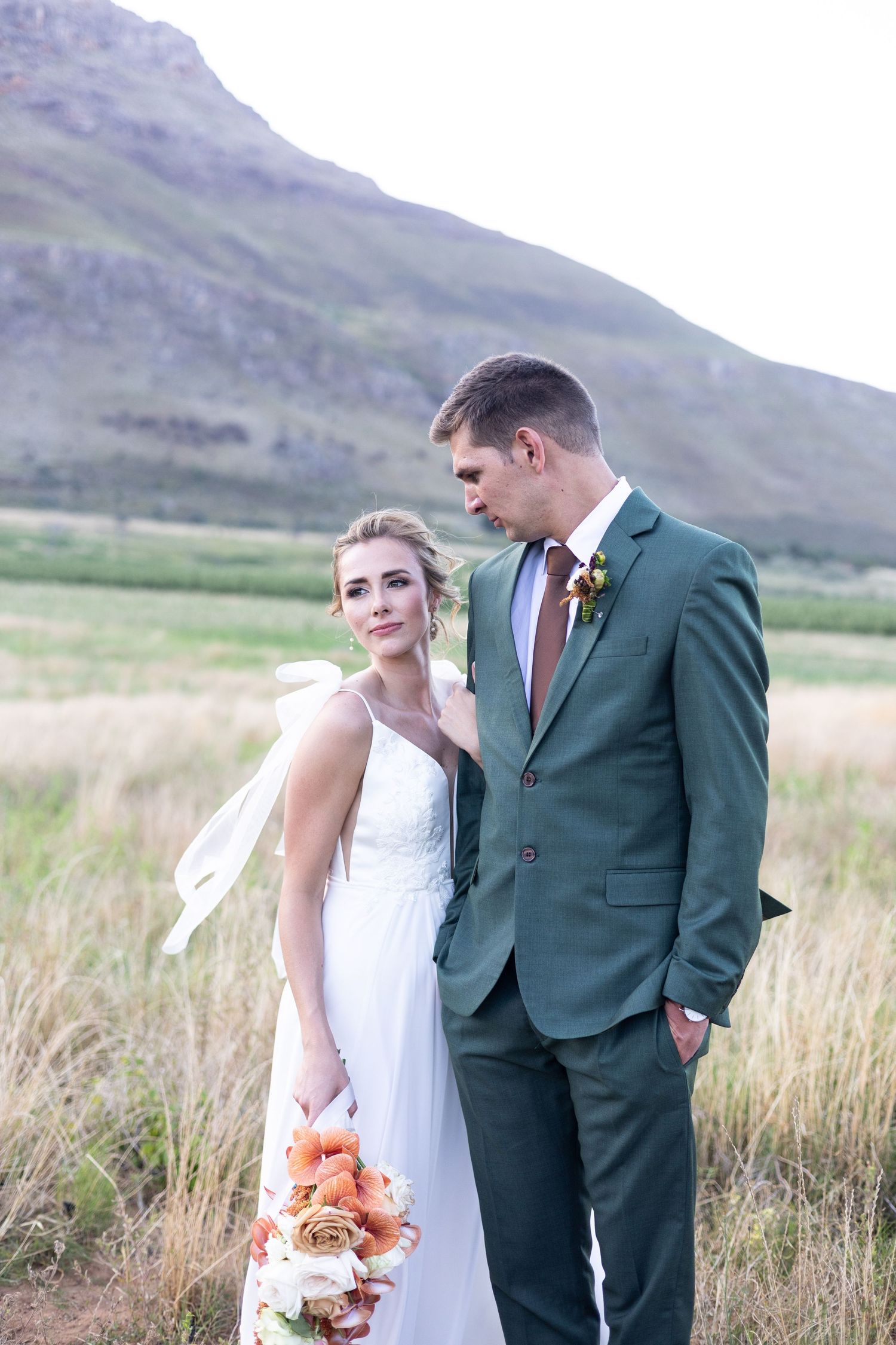 A couple in wedding attire poses in a mountain meadow with tall grass, holding a peach and white floral bouquet.