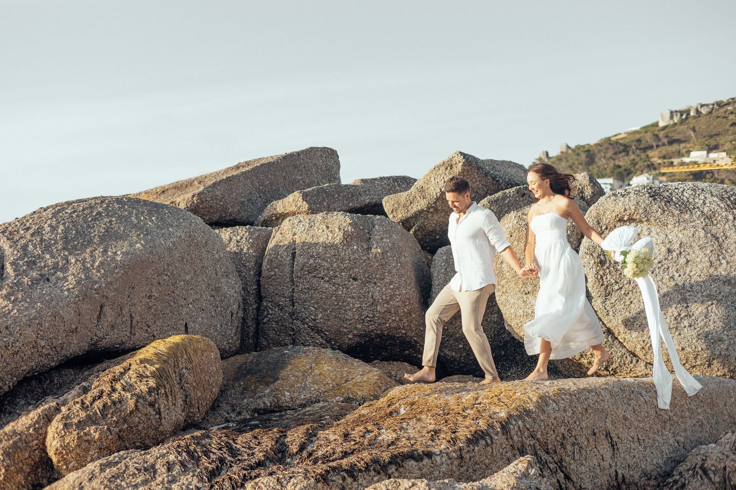 Couple climbs rocky coastal formation in formal white attire.