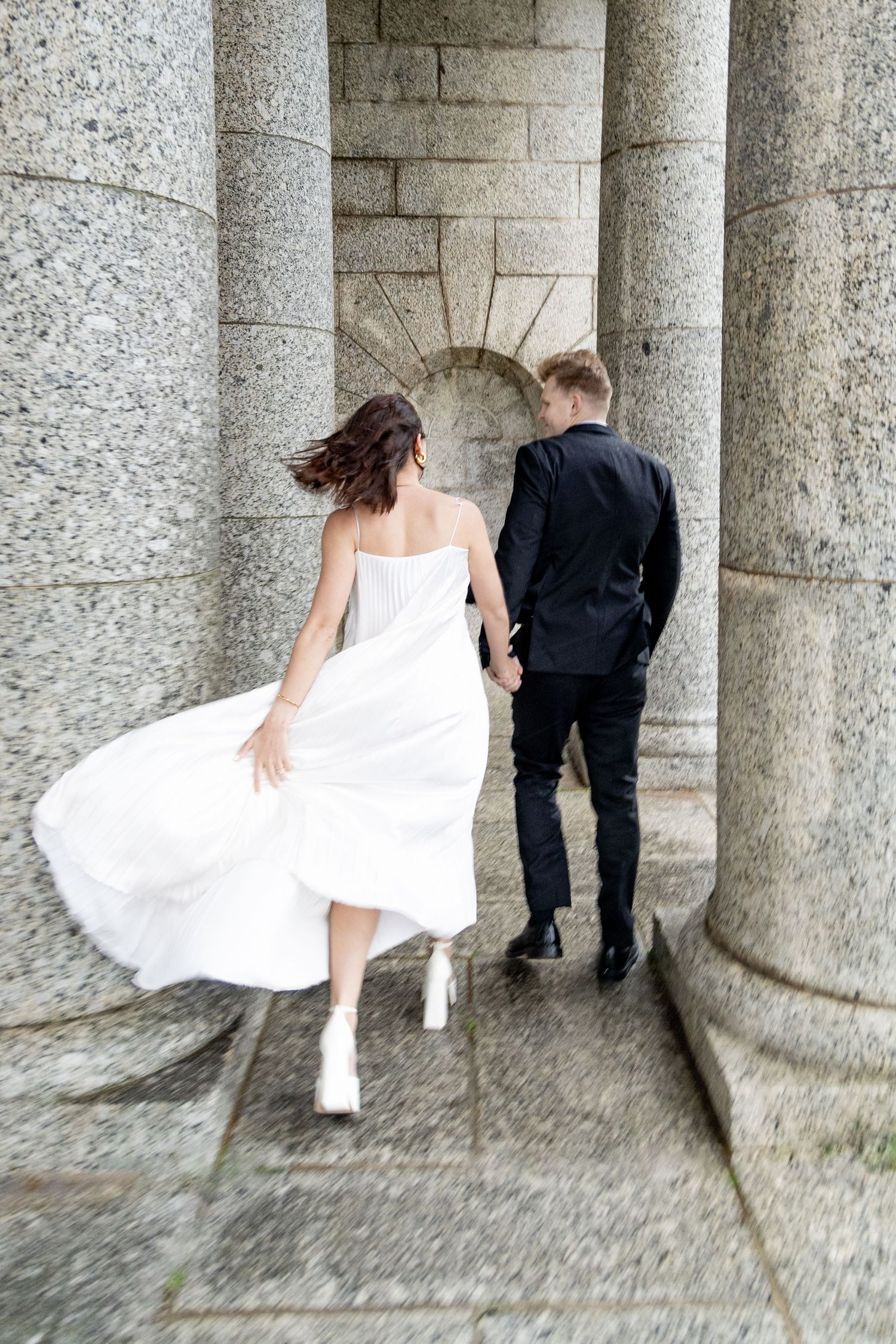 Bride in flowing white dress and groom in black suit walk between classical stone columns.