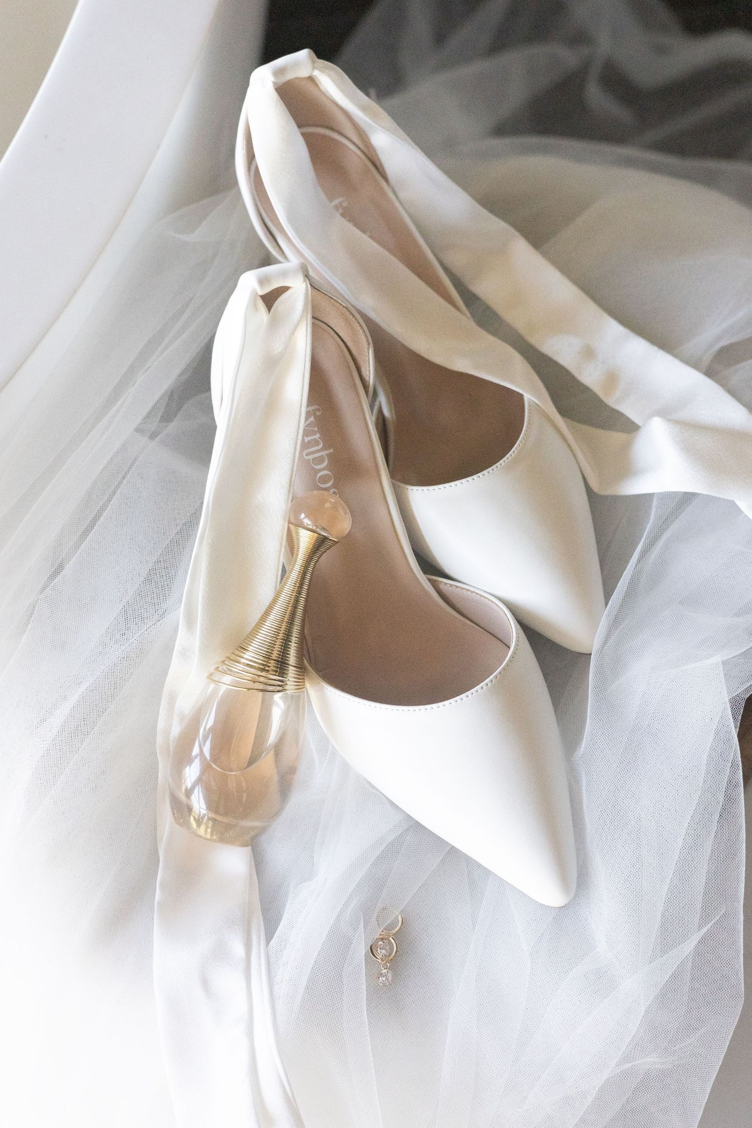 Elegant white satin bridal shoes with pointed toes displayed on soft white tulle fabric with a perfume bottle.