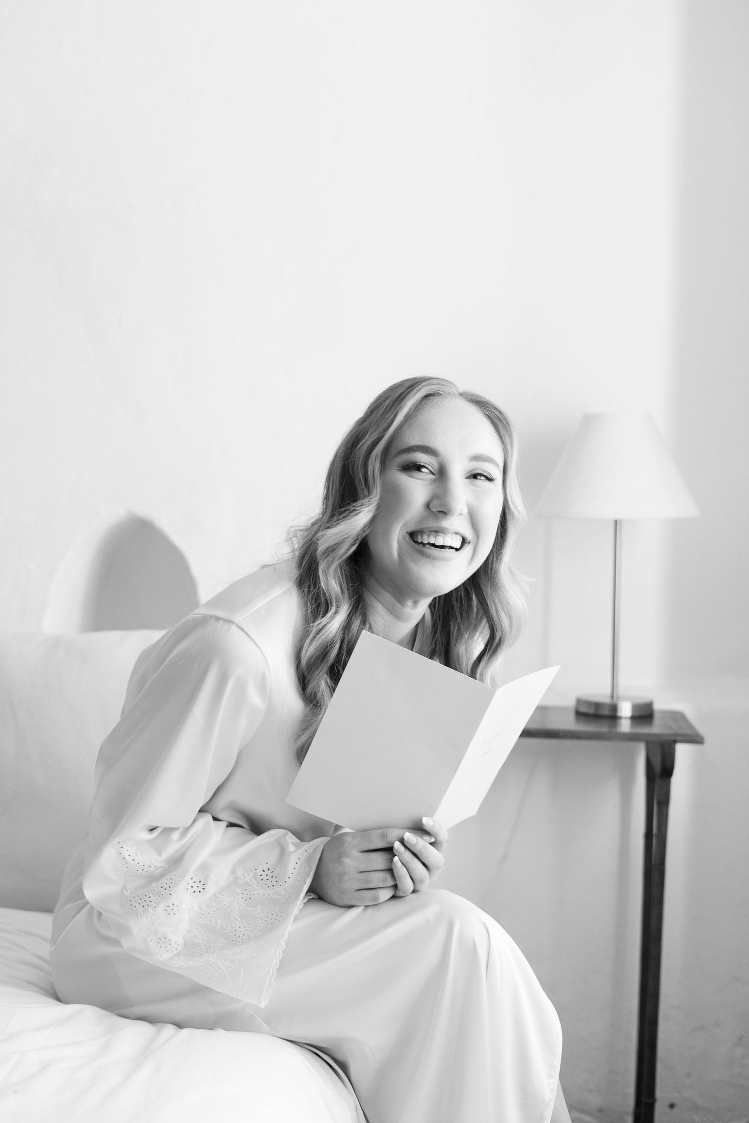 Black and white portrait of person in white robe reading on bed with natural window light.
