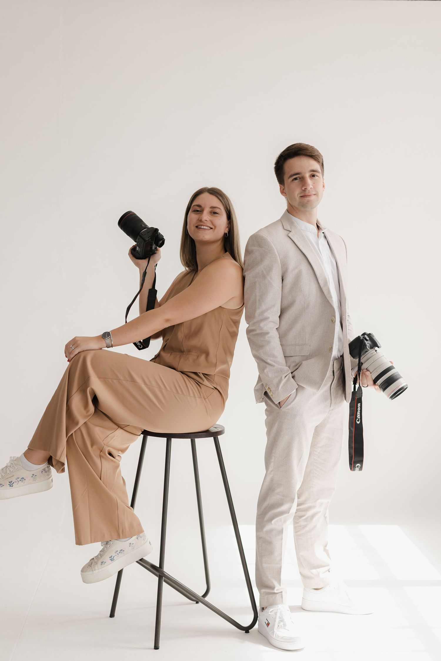 Two photographers pose with professional cameras while wearing coordinated neutral outfits in a minimalist studio setting.