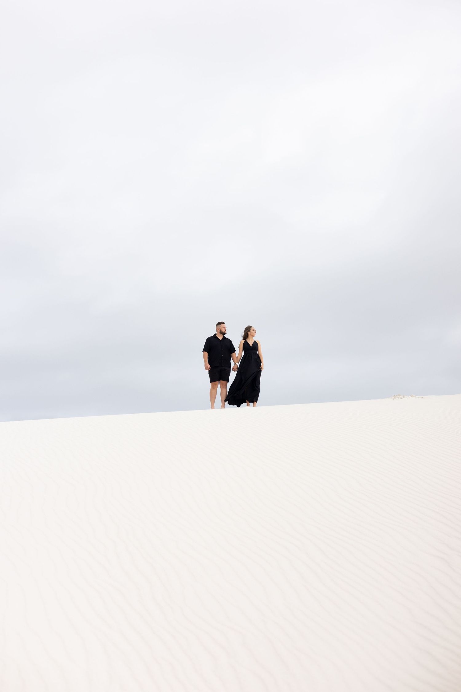 Two figures stand atop a stark white sand dune against an overcast sky in a minimalist desert landscape.