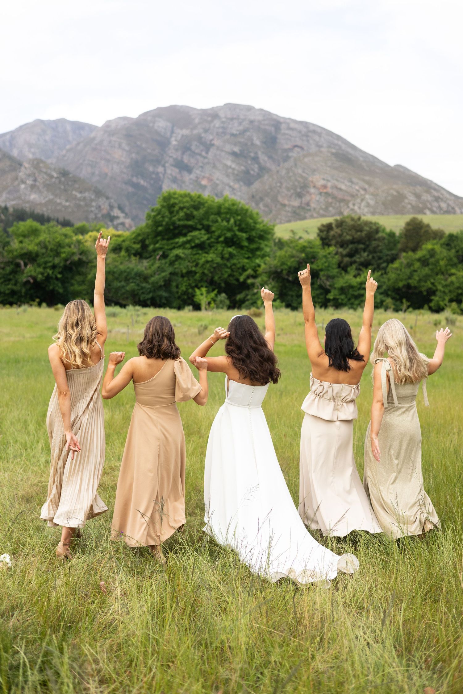 Women in neutral dresses celebrate with raised arms in a grassy field with mountain backdrop.