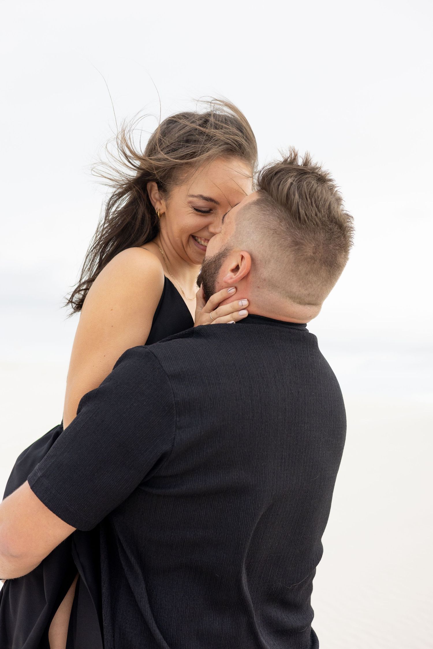 Intimate moment between couple in black attire on beach with wind-blown hair.