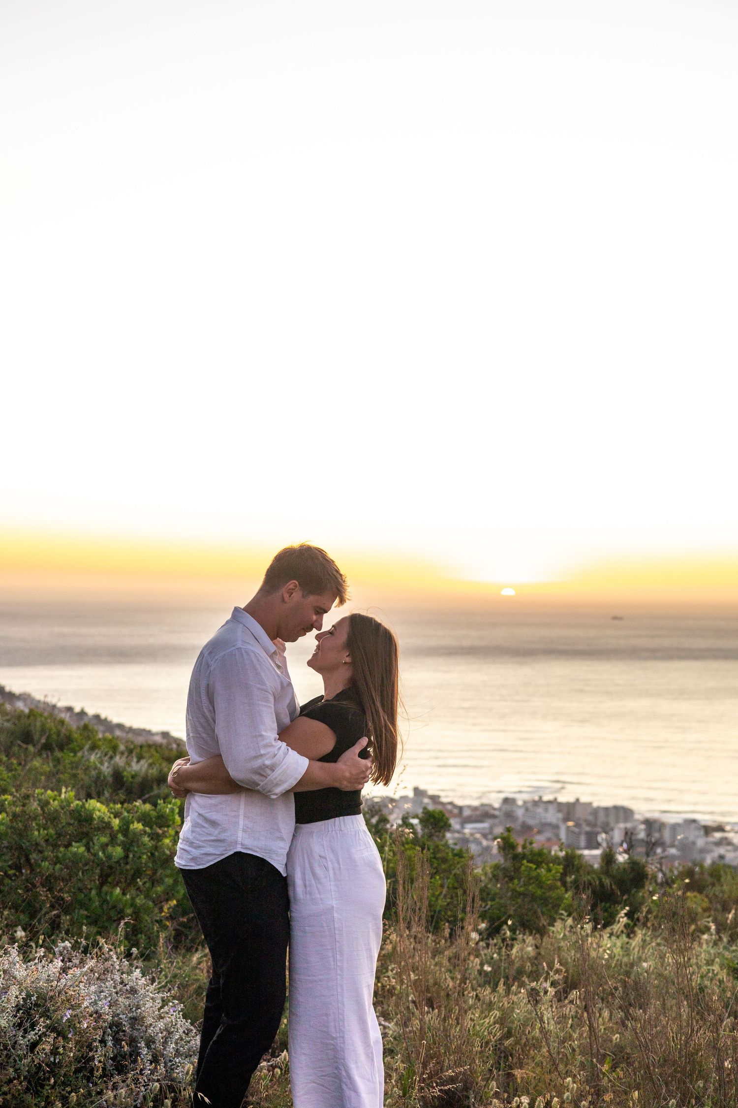 Romantic couple embraces on hillside overlooking coastal city during golden sunset.