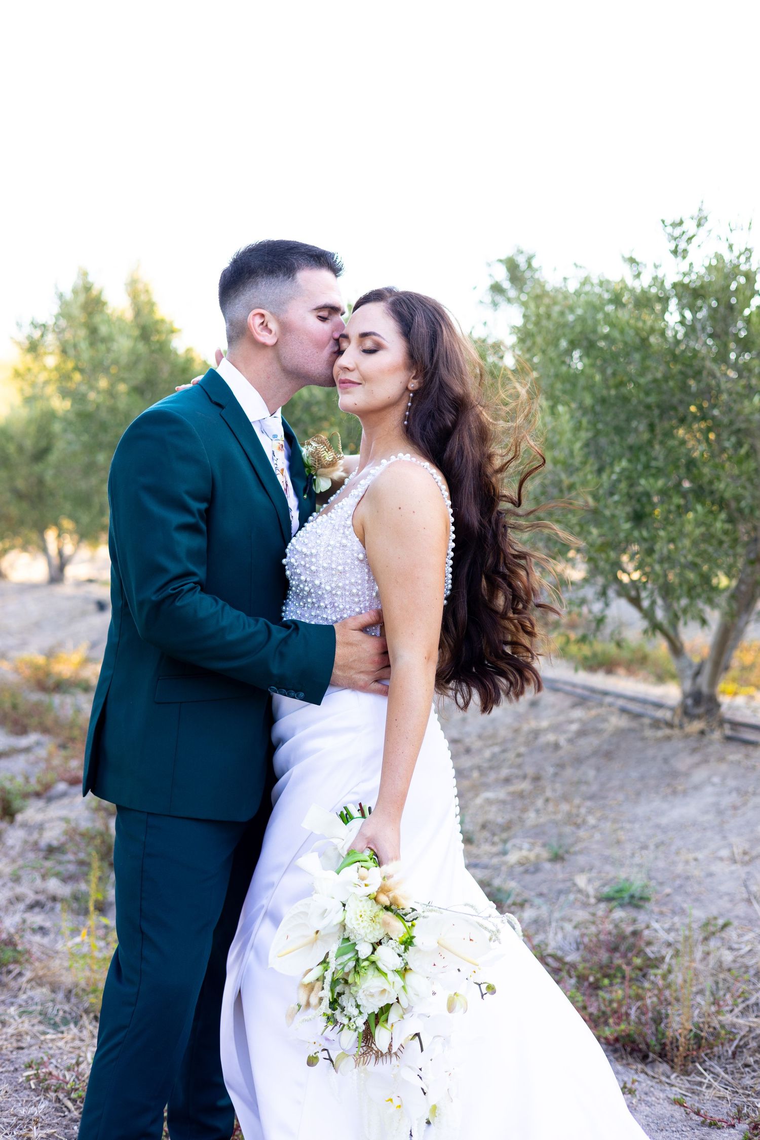 Romantic outdoor wedding photo showing couple embracing in a white dress and dark suit among olive trees with a soft bouquet.
