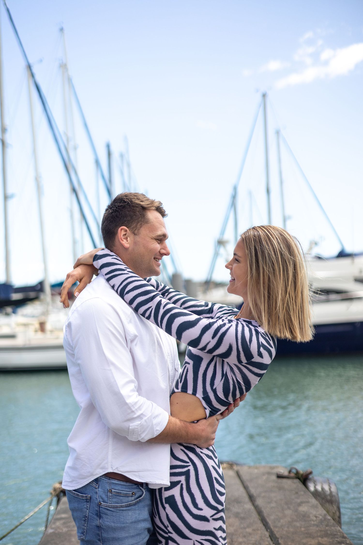 A couple embraces playfully by the marina with sailboats in the background.