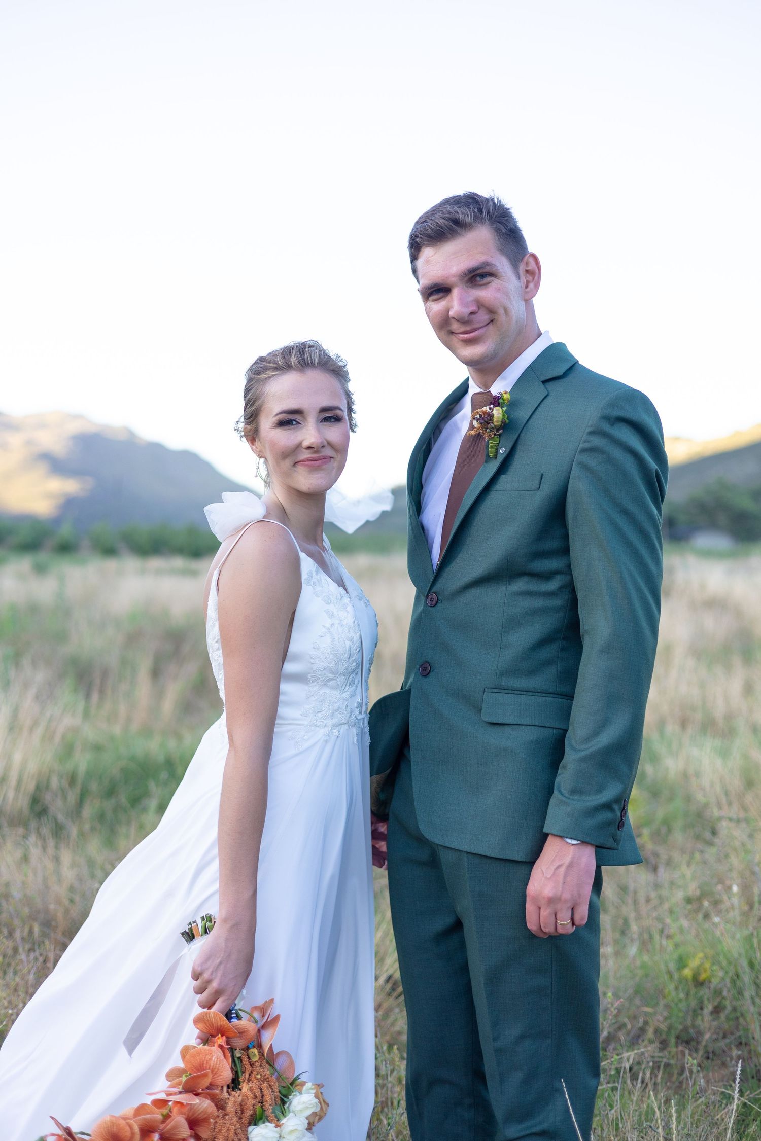 A bride in a white dress and groom in green suit stand together in a mountain field at sunset.