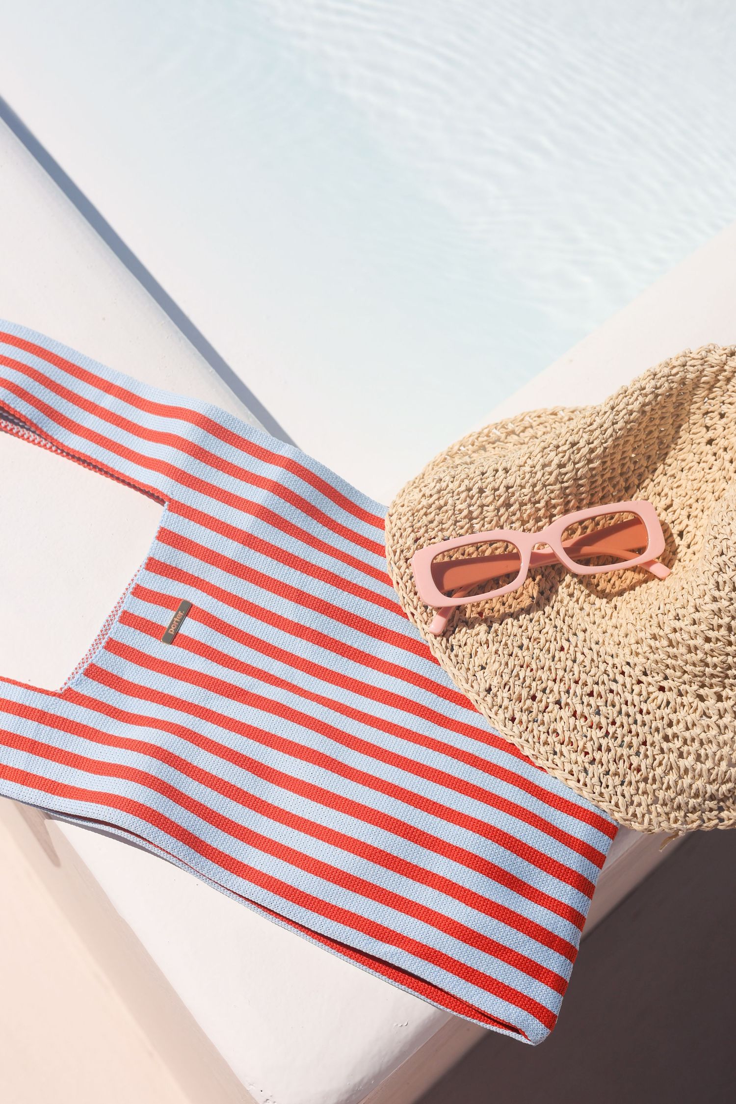 Red and white striped swimsuit with straw sun hat and sunglasses arranged on white surface for summer beach styling.
