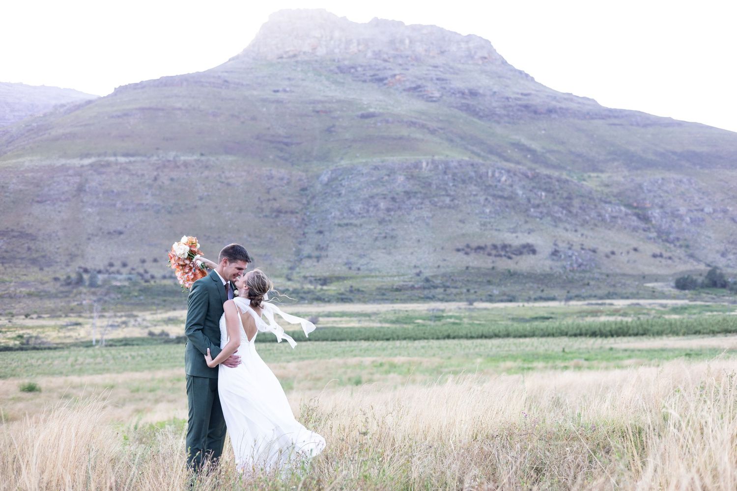 A couple in wedding attire embrace in a grassy field with majestic mountains rising in the misty background.