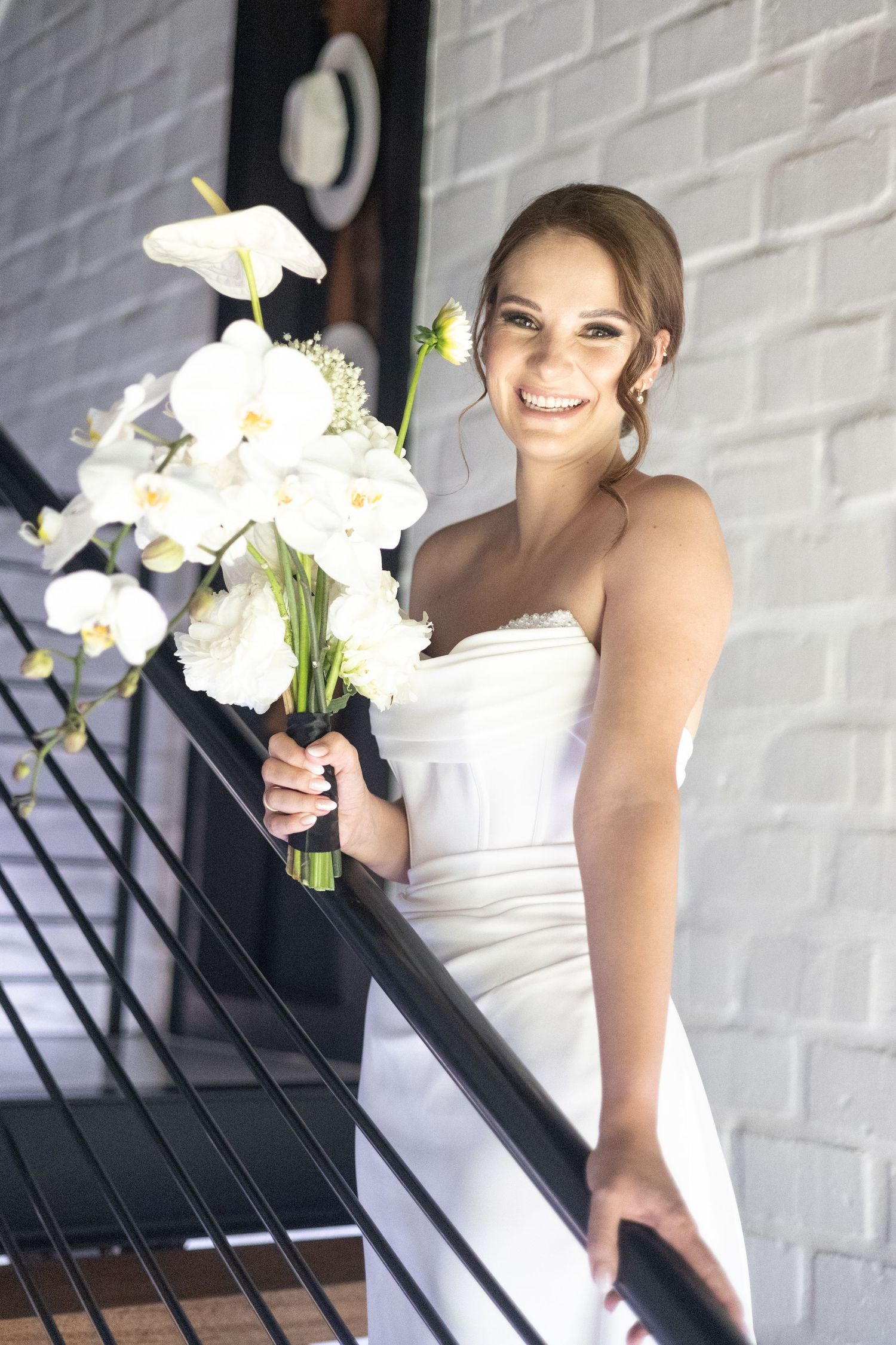 A bride in a strapless white gown holds a bouquet of white orchids and flowers while standing on stairs by a brick wall.