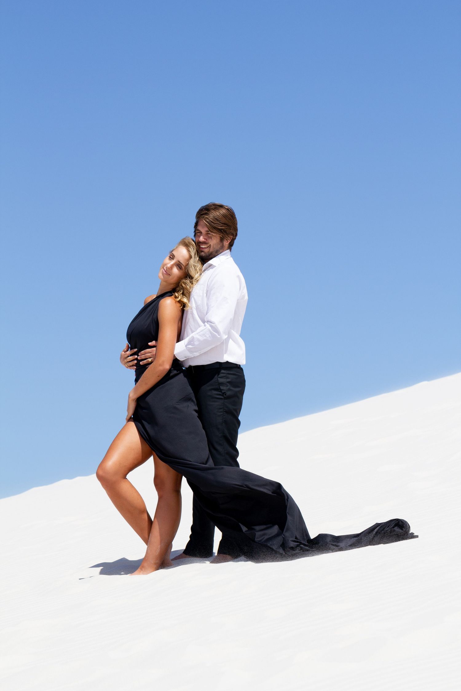 Elegant couple stands together on bright white sand dunes against clear blue sky.