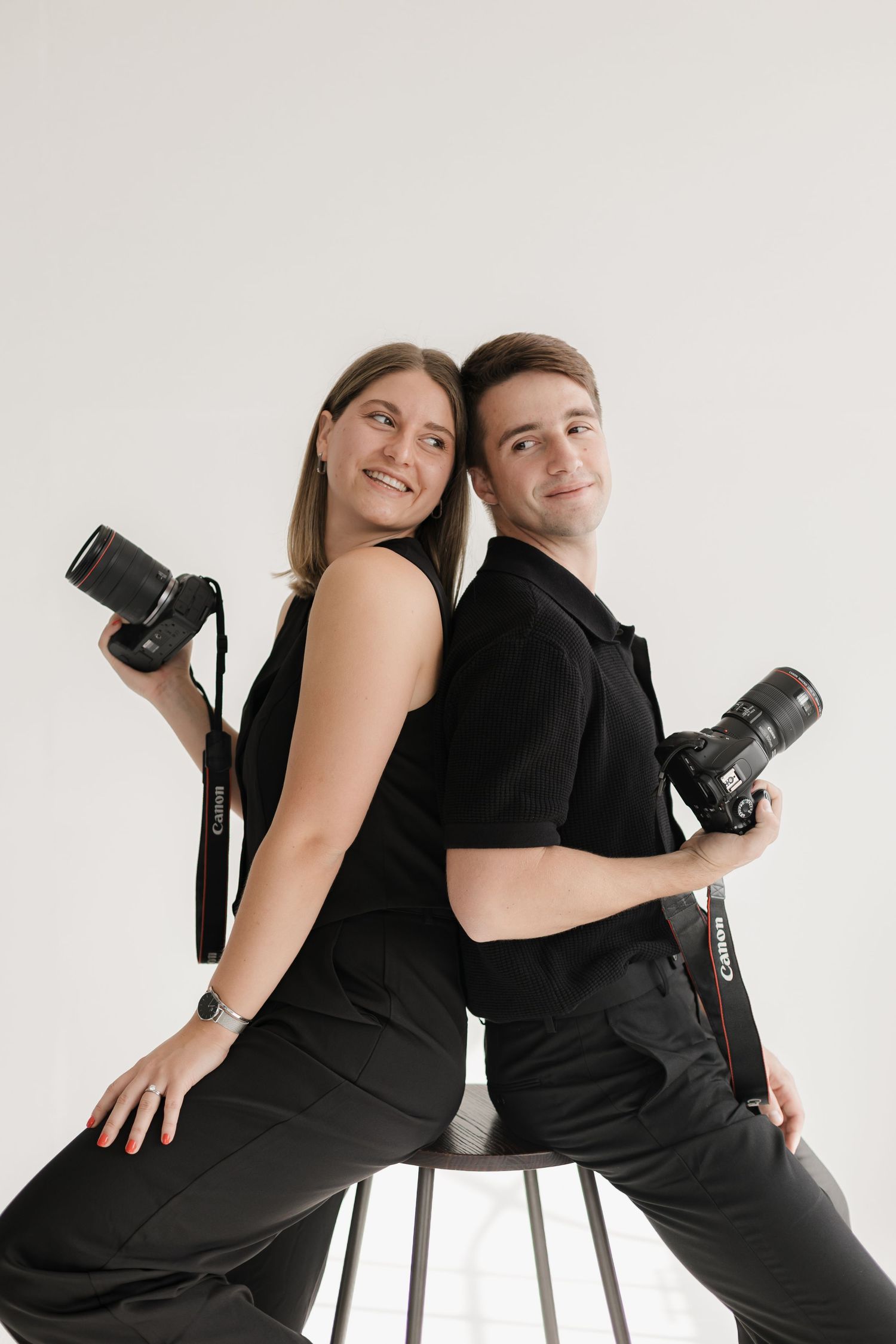 Two photographers in matching black outfits pose back-to-back on a stool with their cameras.