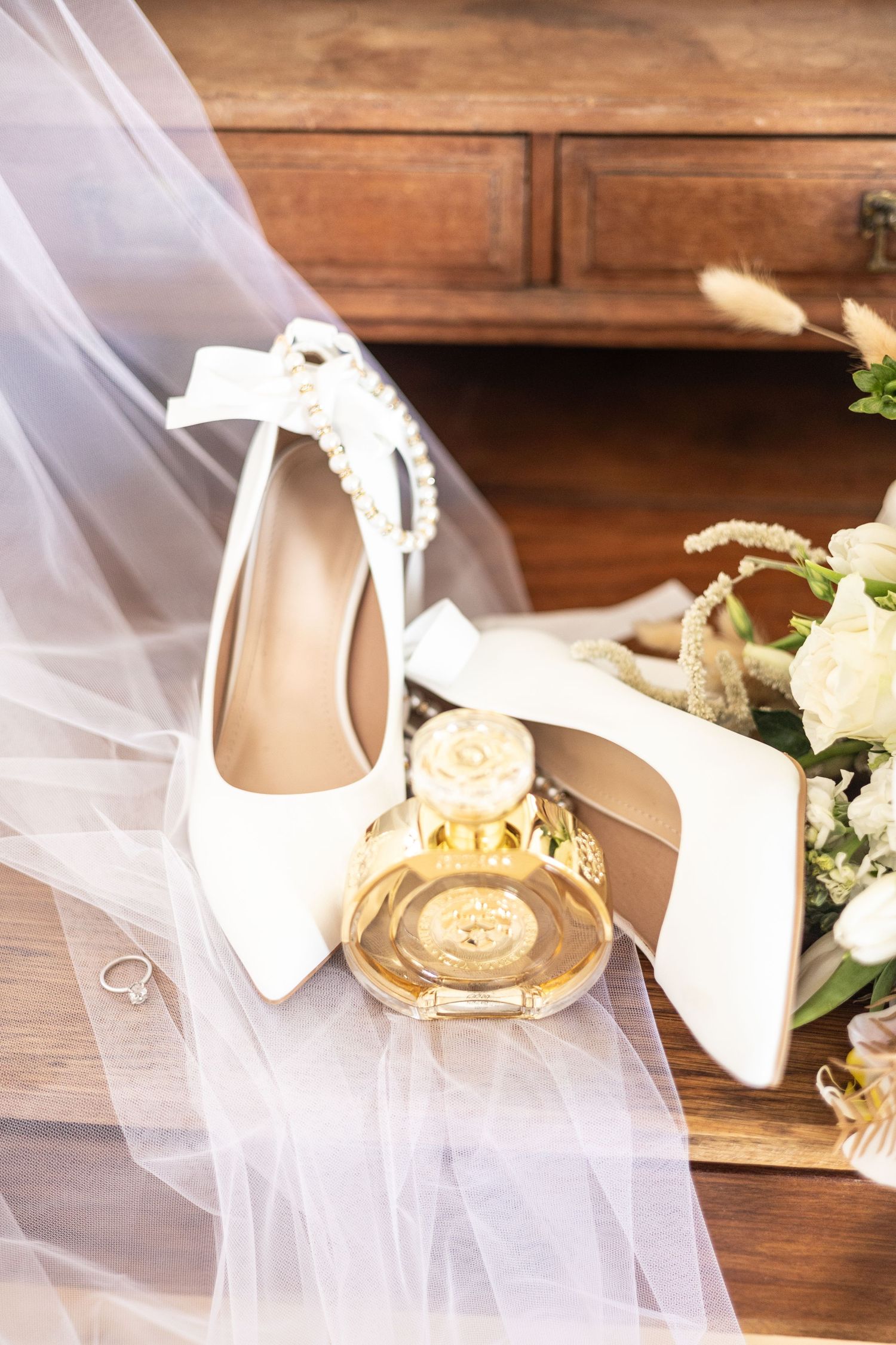 White wedding heels with bridal veil, perfume bottle and floral arrangements on wooden surface.